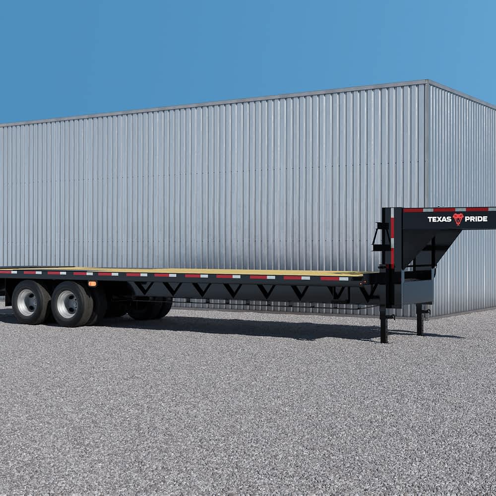 A long, flatbed Texas Pride trailer with multiple axles and a wooden deck is parked on a gravel surface in front of a large, light gray metal building under a clear blue sky. The "TEXAS PRIDE" logo with the longhorn skull is visible on the side of the trailer.