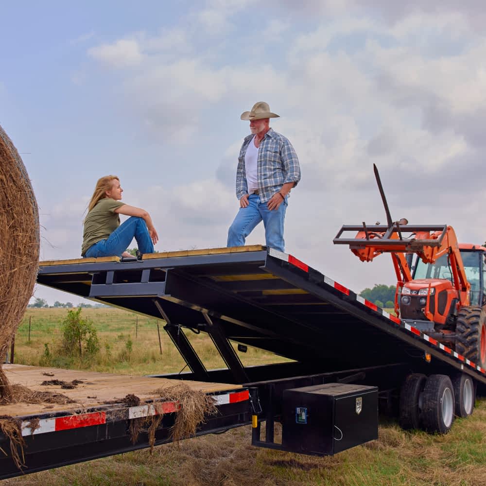 A man in a cowboy hat and a woman are standing and sitting on a black Texas Pride gravity tilt trailer with a hydraulic lift bed. An orange tractor is partially visible on the trailer bed, and a large hay bale sits on the ground next to it in a rural setting under a cloudy sky.