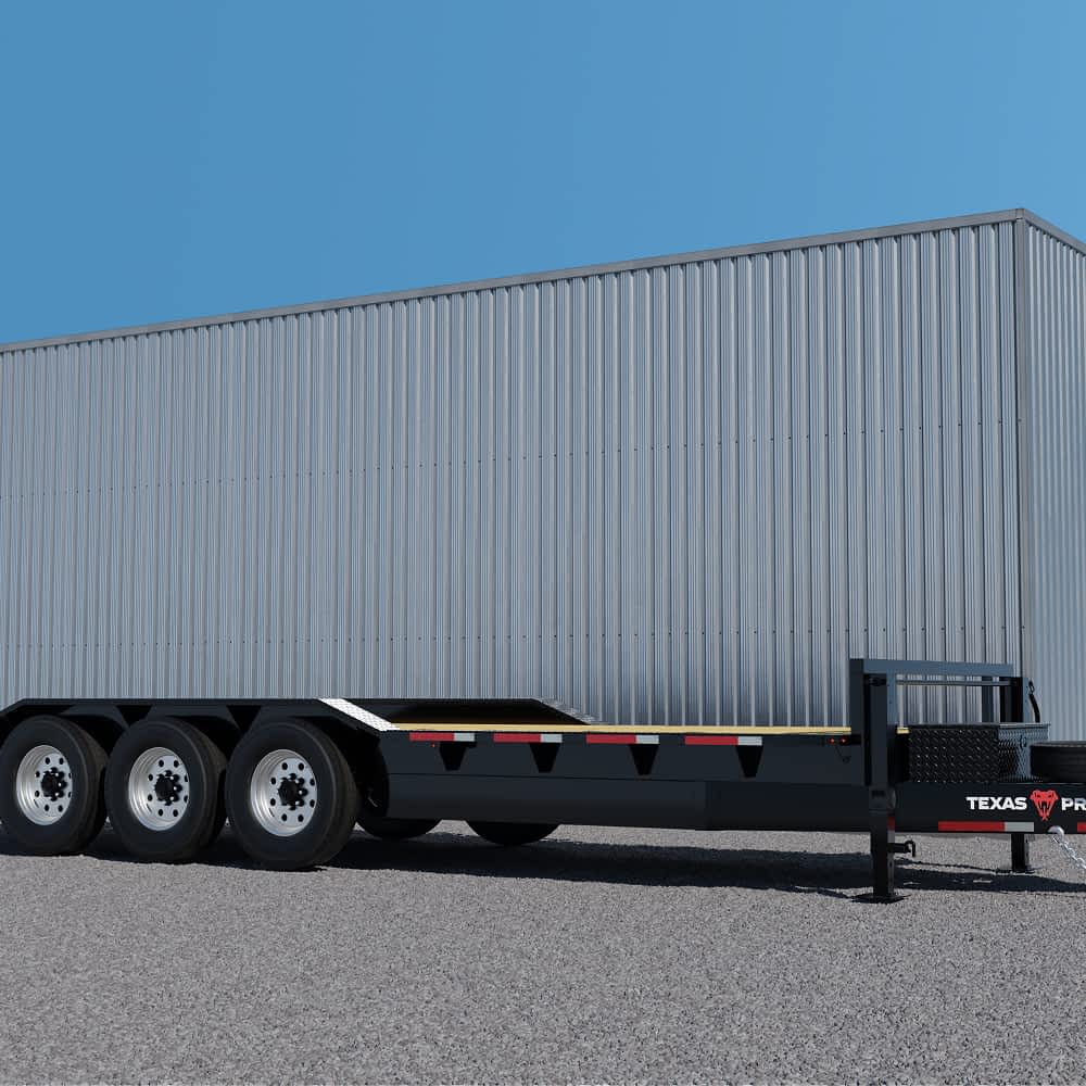 A long, black Texas Pride flatbed lowboy trailer with a wooden deck and multiple axles is parked on a gravel surface in front of a large, light gray metal building under a clear blue sky. The "TEXAS PRIDE" logo is visible on the side.