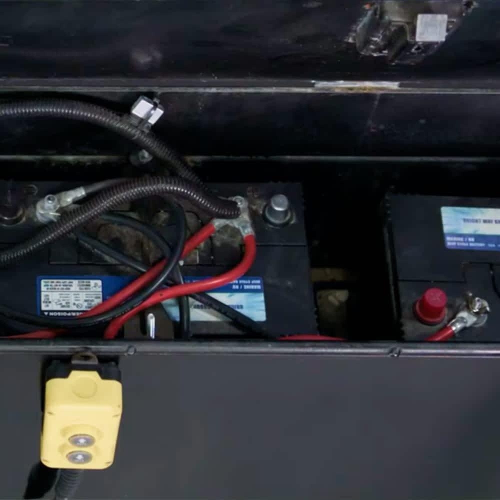 Inside a compartment, on a Texas Pride trailer, are two black batteries with red and black cables connected to their terminals. A yellow control pendant with two buttons is visible hanging below the compartment.