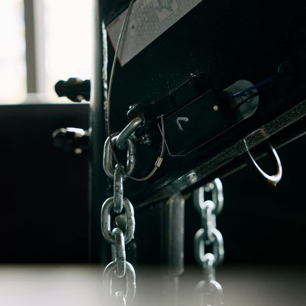 A close-up shows heavy-duty silver chains attached to the underside of a black Texas Pride gooseneck dump trailer. The chains are connected with sturdy hardware, suggesting a secure tie-down system.