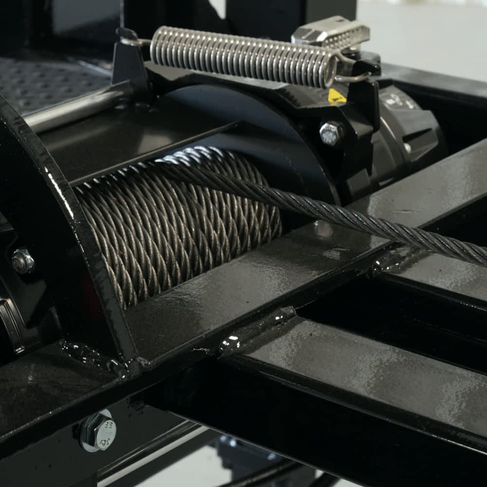 A close-up shows the steel cable spooled around a black winch mechanism, part of a Texas Pride roll-off dumpster trailer. A tension spring and other mechanical components are also visible.