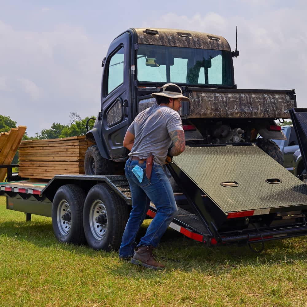 A man wearing a wide-brimmed hat, a gray t-shirt, and jeans is lowering the ramp of a black Texas Pride lowboy trailer. A camouflage-patterned UTV is partially loaded on the trailer, and stacks of lumber are secured on the front section of the trailer bed in a grassy outdoor setting.