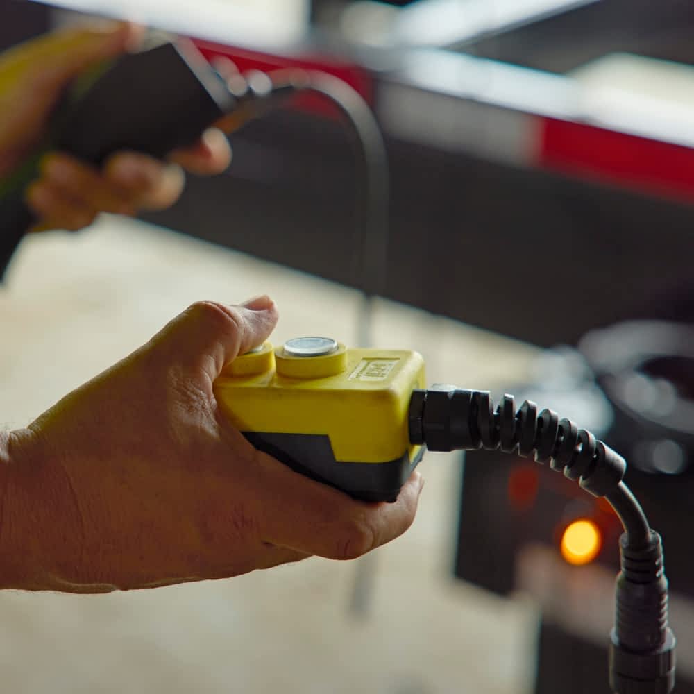 A hand holds a yellow and black control box with buttons, connected by a coiled black cable, used to operate a feature on a Texas Pride roll-off trailer, such as a hydraulic lift. Part of the black trailer with a red reflective strip is visible in the blurred background.