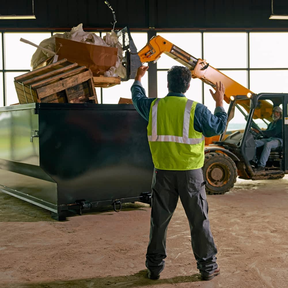 Two workers are using a yellow telescopic handler to lift and dump debris into a large, open-top container. One worker in a bright yellow safety vest and gray pants guides the operator of the machinery, who is seated inside the cab. Two workers are using a yellow telescopic handler to lift and dump debris into a large, open-top roll-off dump container. One worker in a bright yellow safety vest and gray pants guides the operator of the machinery, who is seated inside the cab.