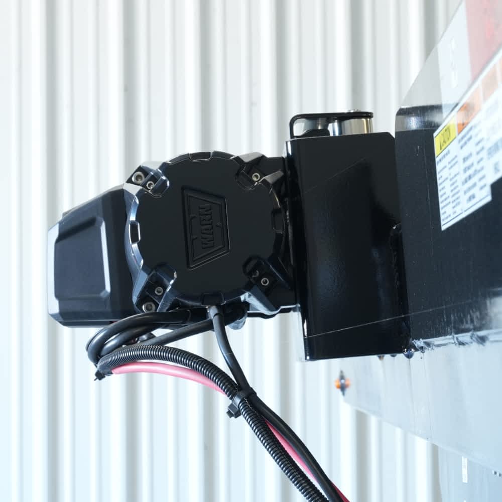 A close-up shows a black 8K WARN winch, mounted on a Texas Pride car hauler trailer, against a light gray corrugated metal background. The winch has various cables extending from it.