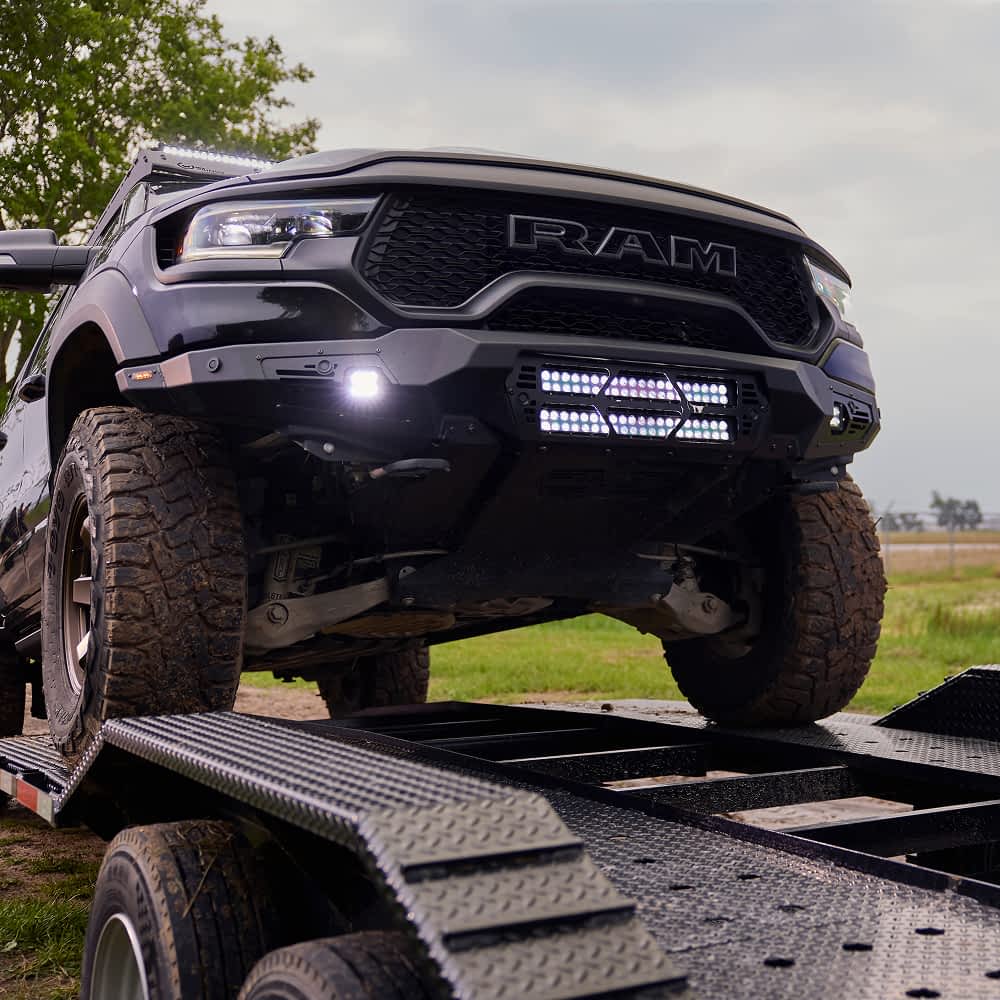 A black RAM truck, a 2024 1500 TRX with an Addictive Desert Designs Stealth Fighter Winch Kit, is being loaded onto a black Texas Pride car hauler trailer with a diamond-plated surface and drive-over fenders. The truck has large, off-road tires and illuminated auxiliary lights.