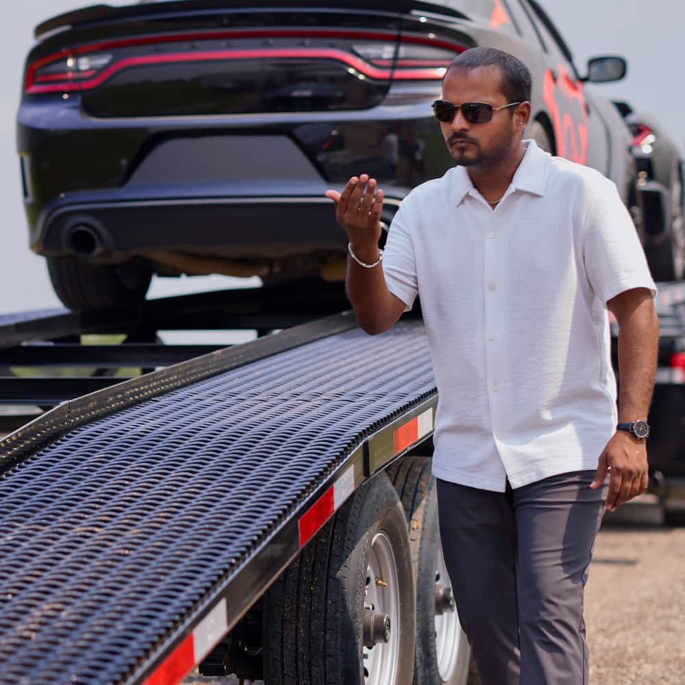 A man wearing sunglasses and a light-colored button-down shirt walks alongside a black Texas Pride car hauler trailer loaded with black Dodge Chargers featuring red accents. The trailer has a grated surface and multiple axles.