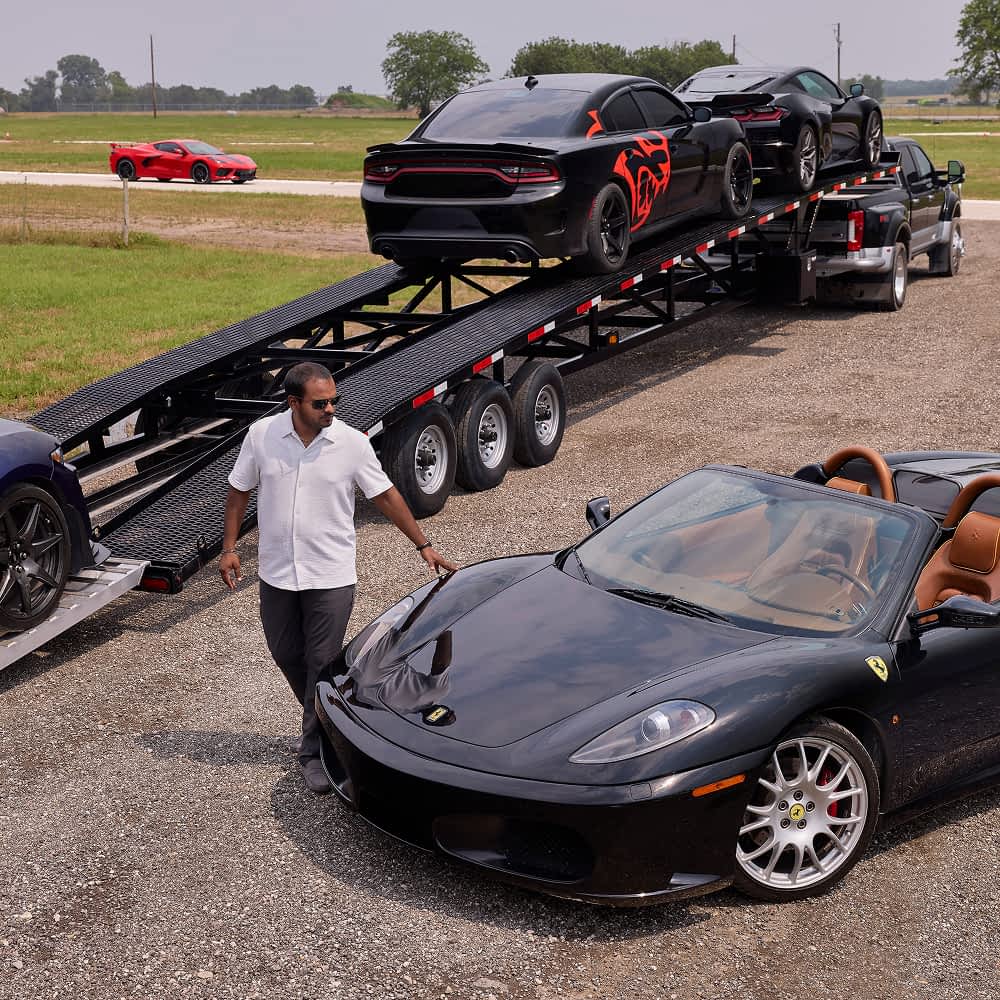 A man in sunglasses and a white shirt walks alongside a black Texas Pride 5-car hauler trailer that is being loaded with multiple vehicles, including black Dodge Chargers with red racing stripes. A black Ferrari Modena convertible with a tan interior is parked in the foreground, and a red Corvette is visible in the distance on a paved rack track next to a grassy field.