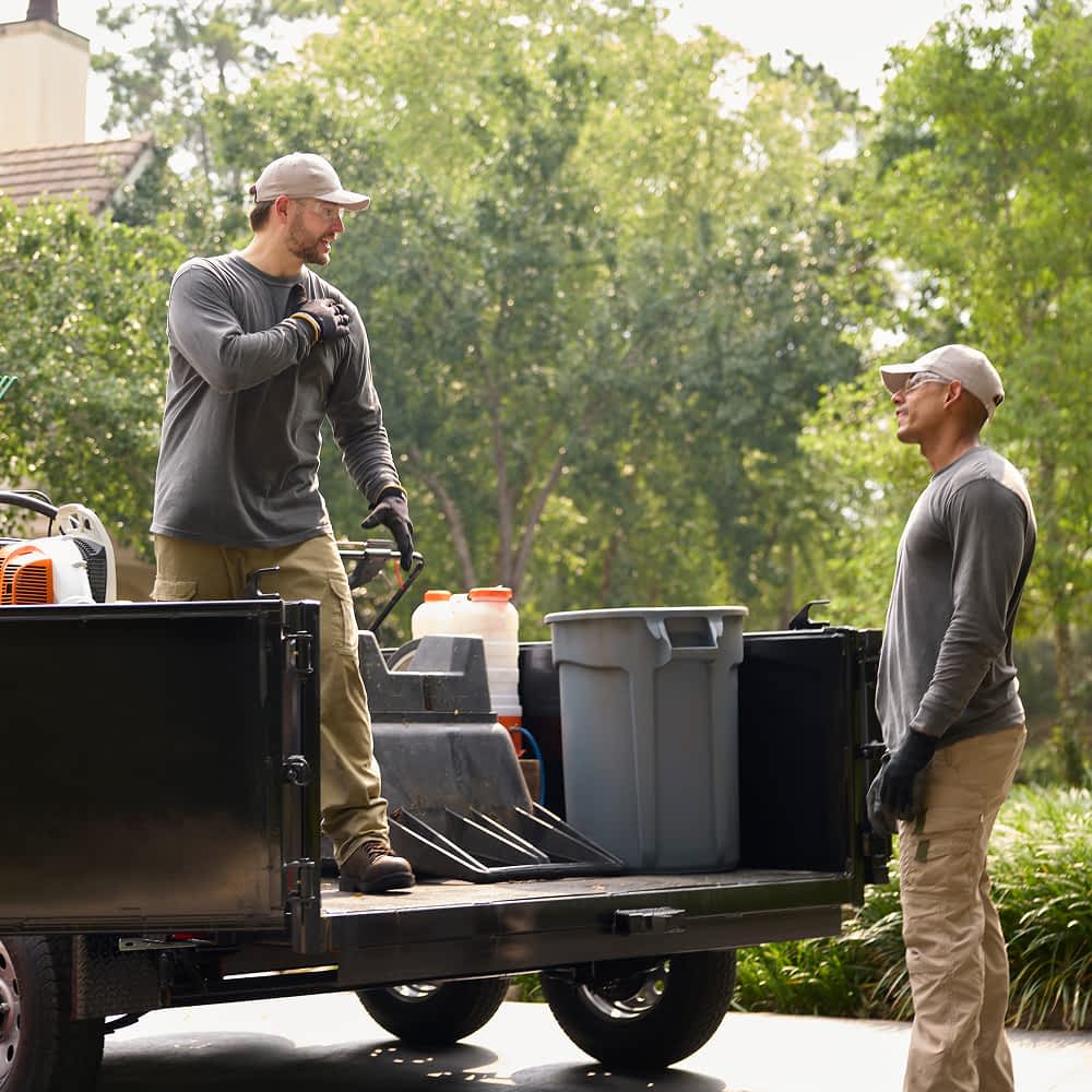 Two workers in outdoor work attire load equipment onto a heavy-duty Texas Pride dump trailer. One stands on the trailer bed, securing tools and containers, while the other listens attentively beside the trailer.