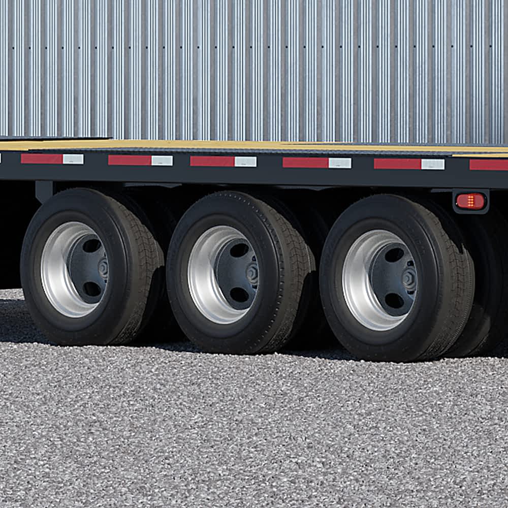 A close-up shows the dual wheel triple axles with silver wheels and black tires of a Texas Pride flatbed trailer parked on gray gravel. Red and white reflective tape runs along the side of the trailer above the wheels.