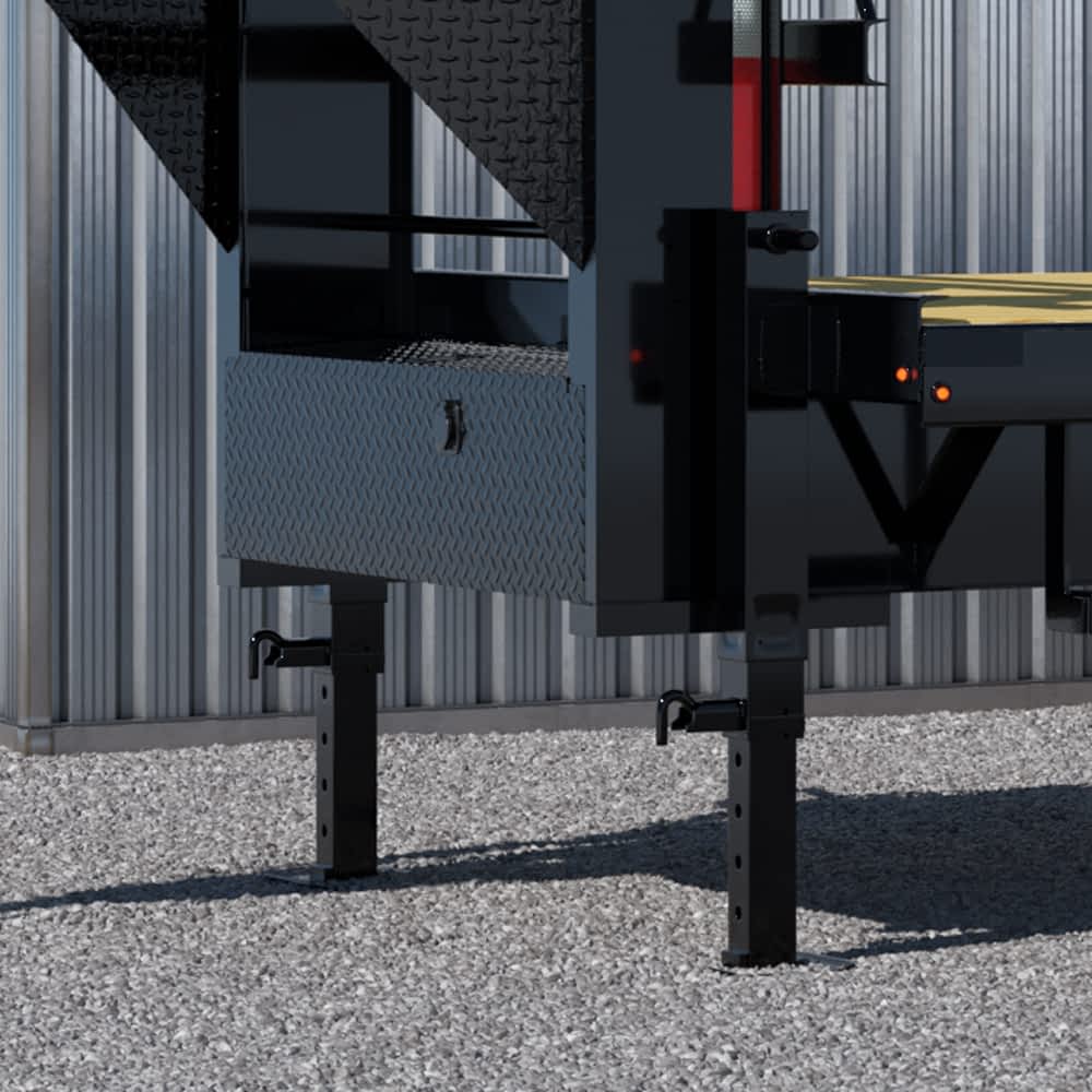 A detailed view shows the adjustable support legs and diamond-plated storage compartment of a black Texas Pride flatbed trailer parked on gravel. The side of a corrugated metal building is visible in the background.
