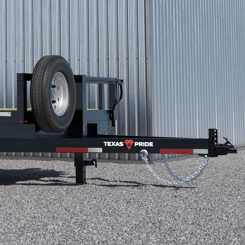 A close-up shows the hitch and side of a black Texas Pride gravity tilt trailer with a spare tire mounted near the front. The trailer features the "TEXAS PRIDE" logo in white letters next to a red longhorn skull graphic, above a red and white reflective safety strip.