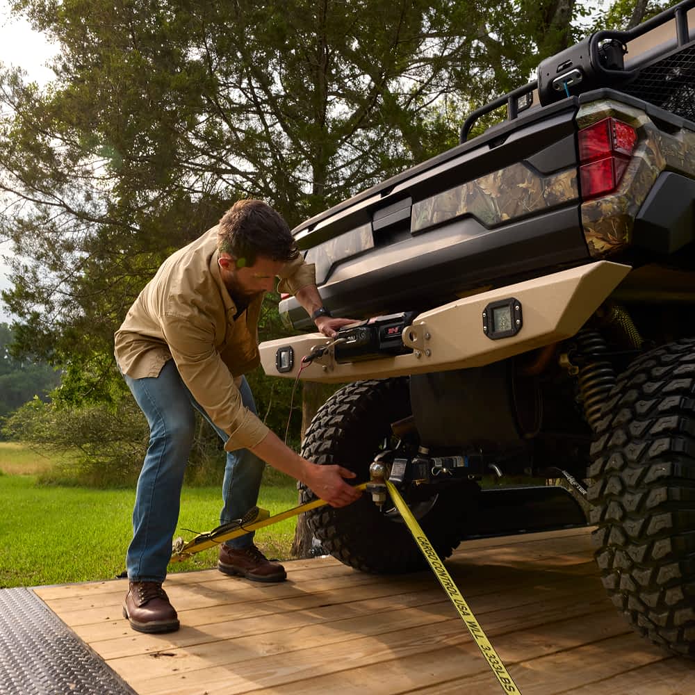 A man in a tan shirt and blue jeans is securing a camouflage-patterned side-by-side utility terrain vehicle (UTV) onto a Texas Pride gravity tilt trailer using yellow ratchet straps. The UTV has large, knobby tires, and the trailer has a wooden deck.