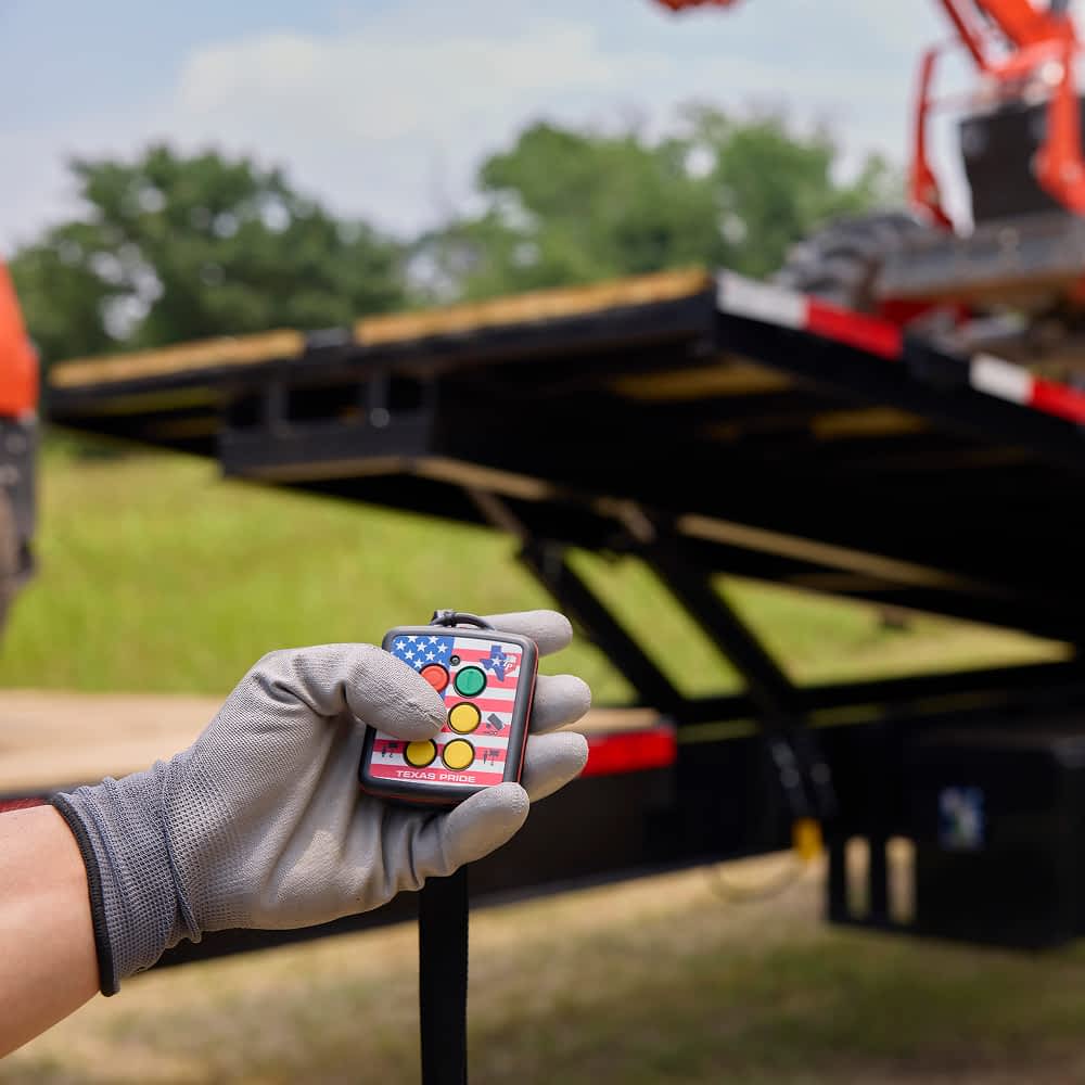 A hand wearing a gray work glove holds a wireless remote control with colorful buttons and a Texas Pride logo, with a black Texas Pride power tilt trailer and a piece of orange machinery visible in the blurred background. The remote likely operates a function of the trailer or machinery.