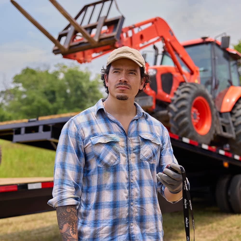 A man wearing a tan baseball cap, a blue and white plaid shirt, and gray gloves stands in front of an orange tractor that is partially loaded onto a black Texas Pride power tilt trailer. The man has a mustache and a tattoo visible on his left forearm.