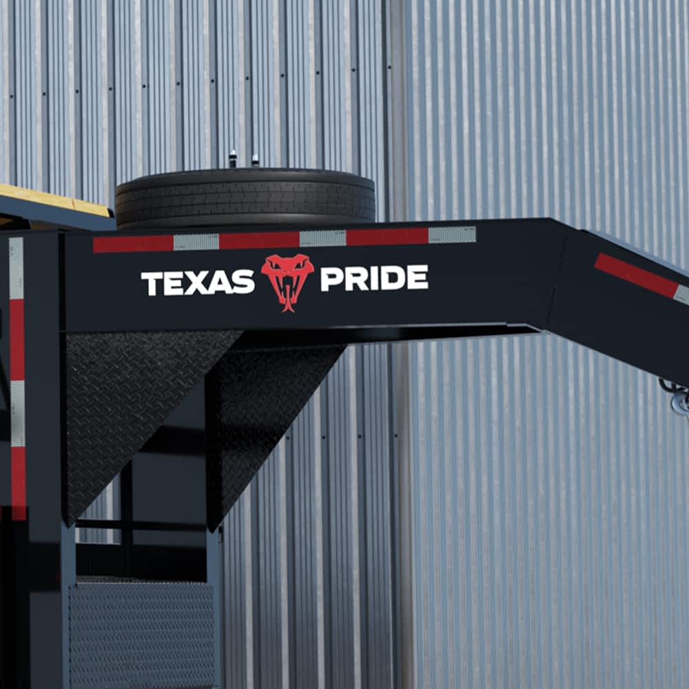A portion of a black Texas Pride power tilt trailer is shown against a corrugated metal wall, highlighting the white "TEXAS" lettering, a red longhorn skull graphic, and the white "PRIDE" lettering. A spare tire is visible mounted on the trailer.