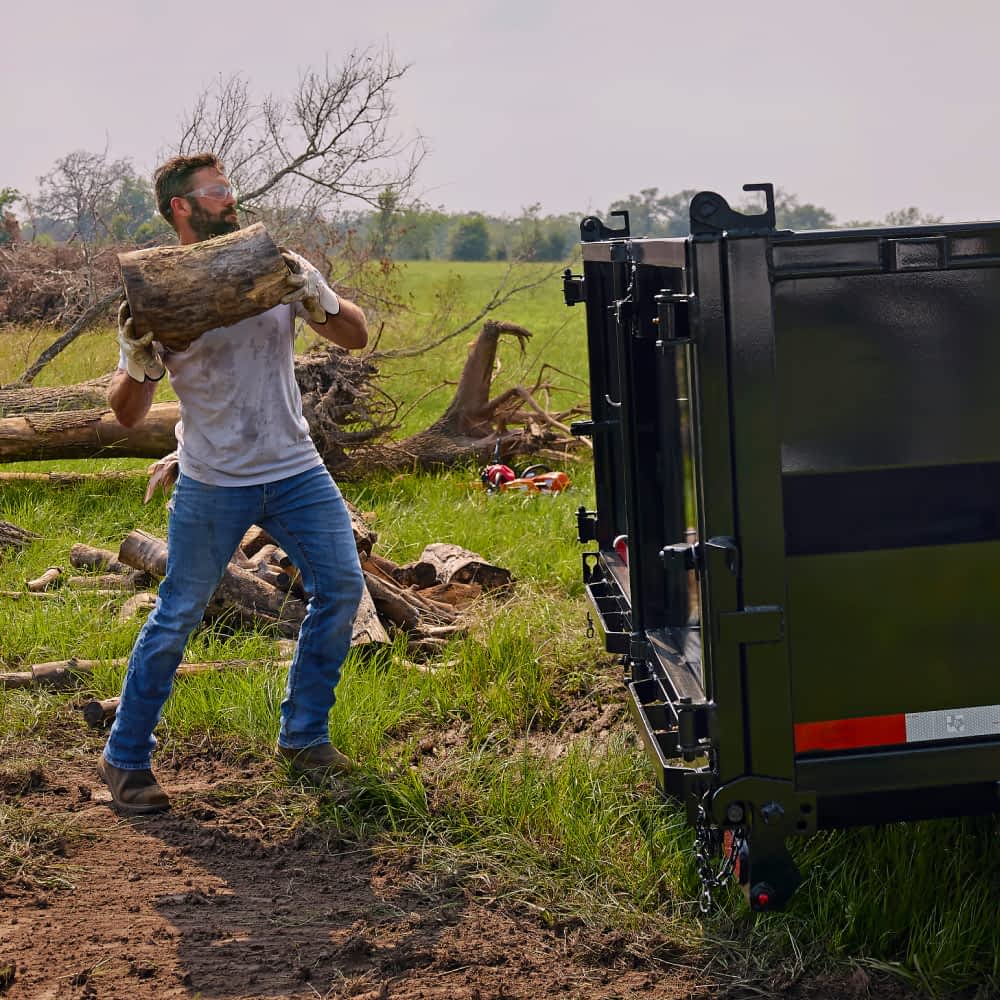 A man with a beard and safety glasses carries a large log towards a black Texas Pride dump trailer with a red reflective strip, parked on a grassy field with fallen tree limbs. The man is wearing a light-colored t-shirt, jeans, and work boots, and appears to be loading the trailer.