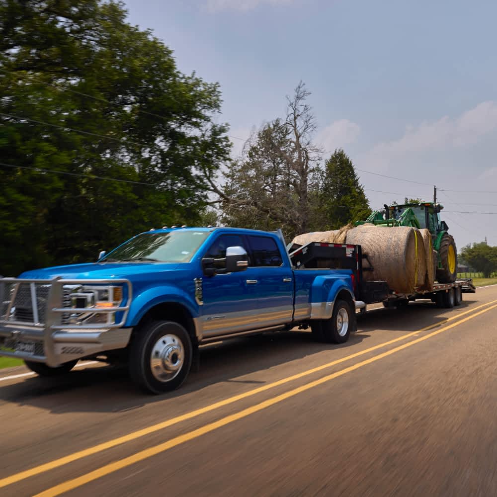 A bright blue Ford Super Duty truck is driving on a road with double yellow lines, towing a Texas Pride gravity tilt trailer. The trailer carries a green John Deere tractor and a large round hay bale wrapped in white netting.