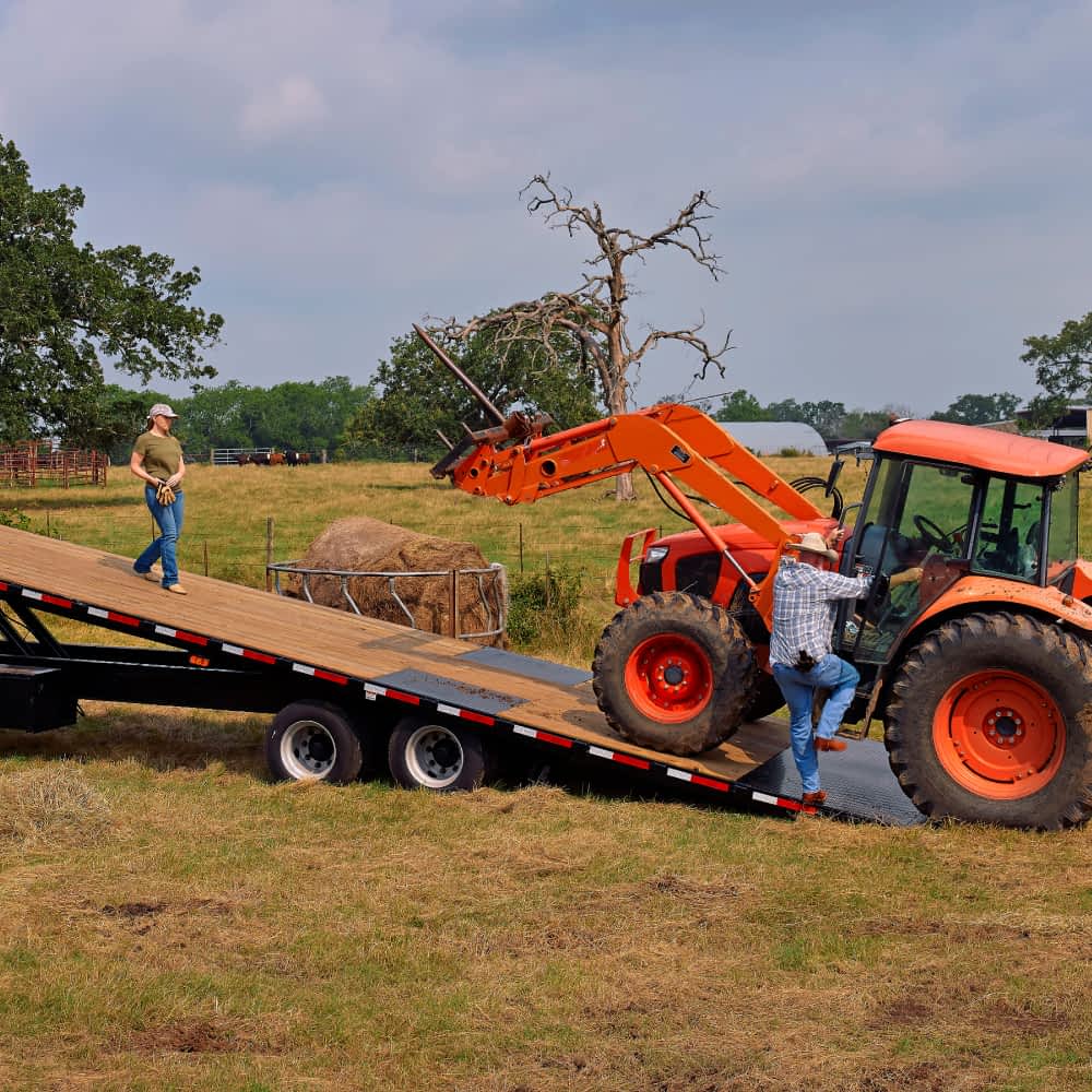 A man in a plaid shirt and jeans is driving an orange tractor with a front-end loader up the ramp of a black Texas Pride power tilt trailer, while a woman in a green shirt and jeans stands on the trailer bed. A round hay bale sits in a field next to the trailer in a rural landscape under a partly cloudy sky.