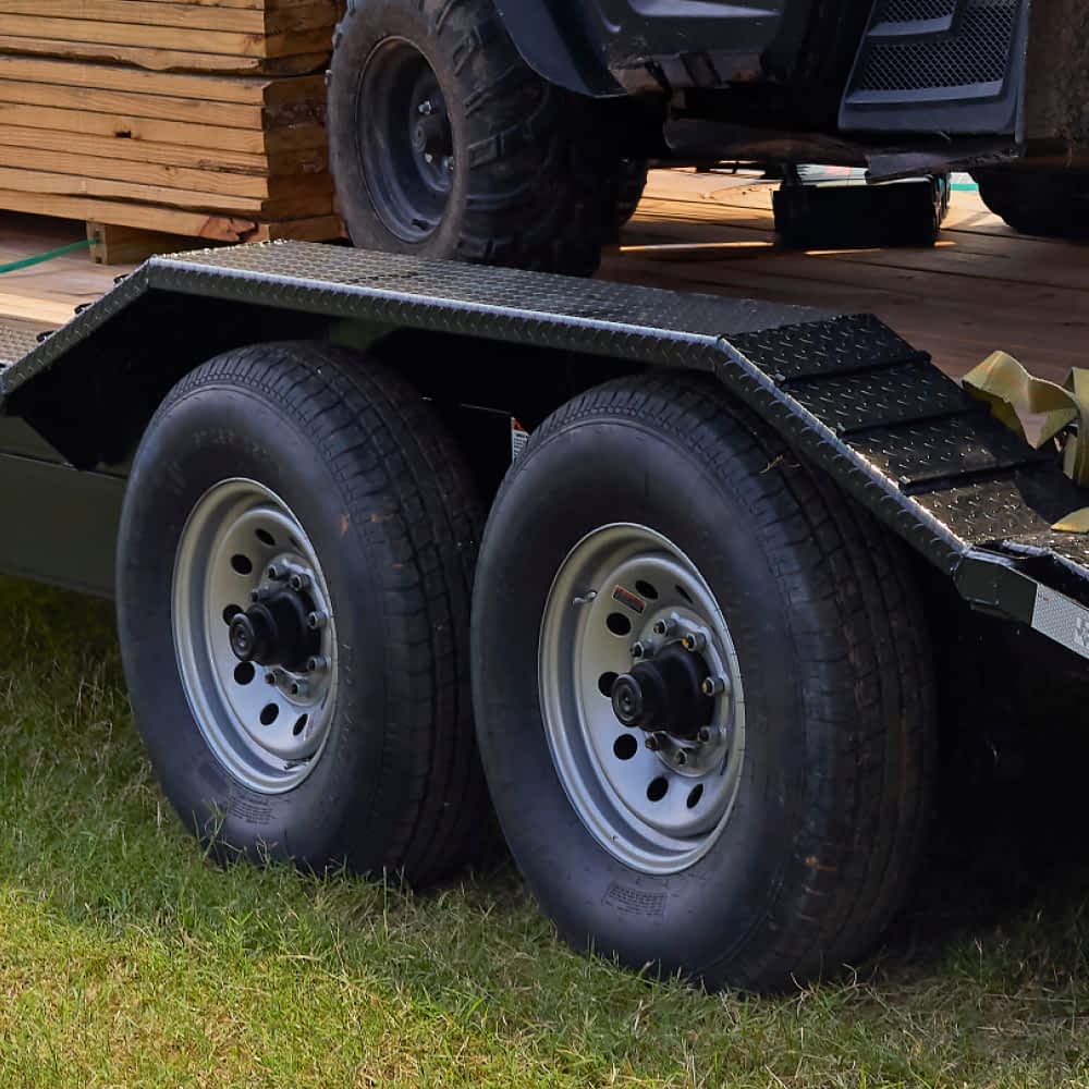 A close-up shows the drive-over fenders above tandem axles with silver wheels and black tires of a black Texas Pride lowboy trailer parked on grass. A stack of lumber and part of a UTV are visible on the trailer bed above the wheels.
