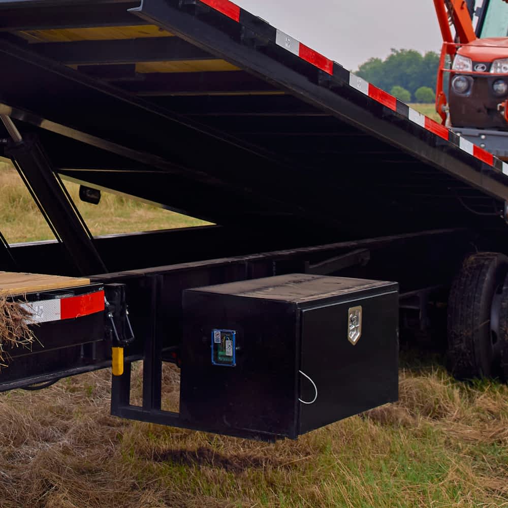 A black toolbox is mounted underneath a black Texas Pride power tilt trailer with a hydraulic lift bed. Part of an orange tractor is visible on the tilted trailer bed in a grassy outdoor setting.