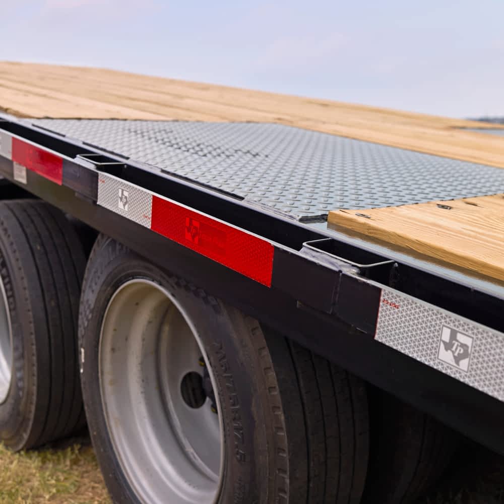 A detailed view shows the wheels, frame, and wooden deck of a Texas Pride power tilt trailer. Red and white reflective tape runs along the side, and a "TP" logo is visible on a reflective marker.