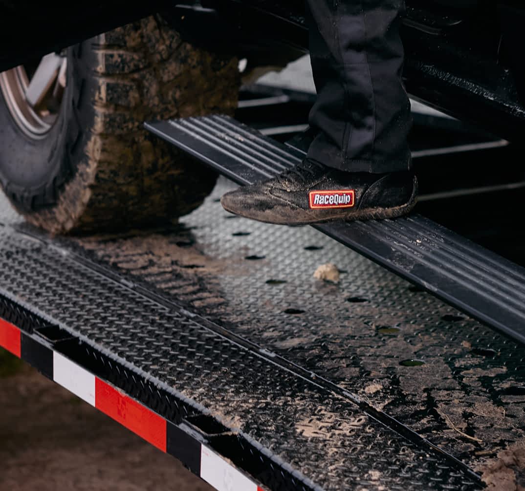 A close-up shows a person's foot, wearing a black shoe with a "RaceQuip" patch, stepping onto the diamond-plated ramp of a black Texas Pride car hauler trailer. A drive-over-fender is shown with white and red reflective tape. A muddy tire of a vehicle being loaded is visible in the background.