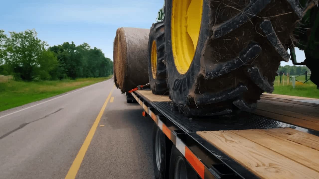 A Texas Pride Trailers flatbed trailer loaded with a hay bale and a green tractor is pulled down a road.