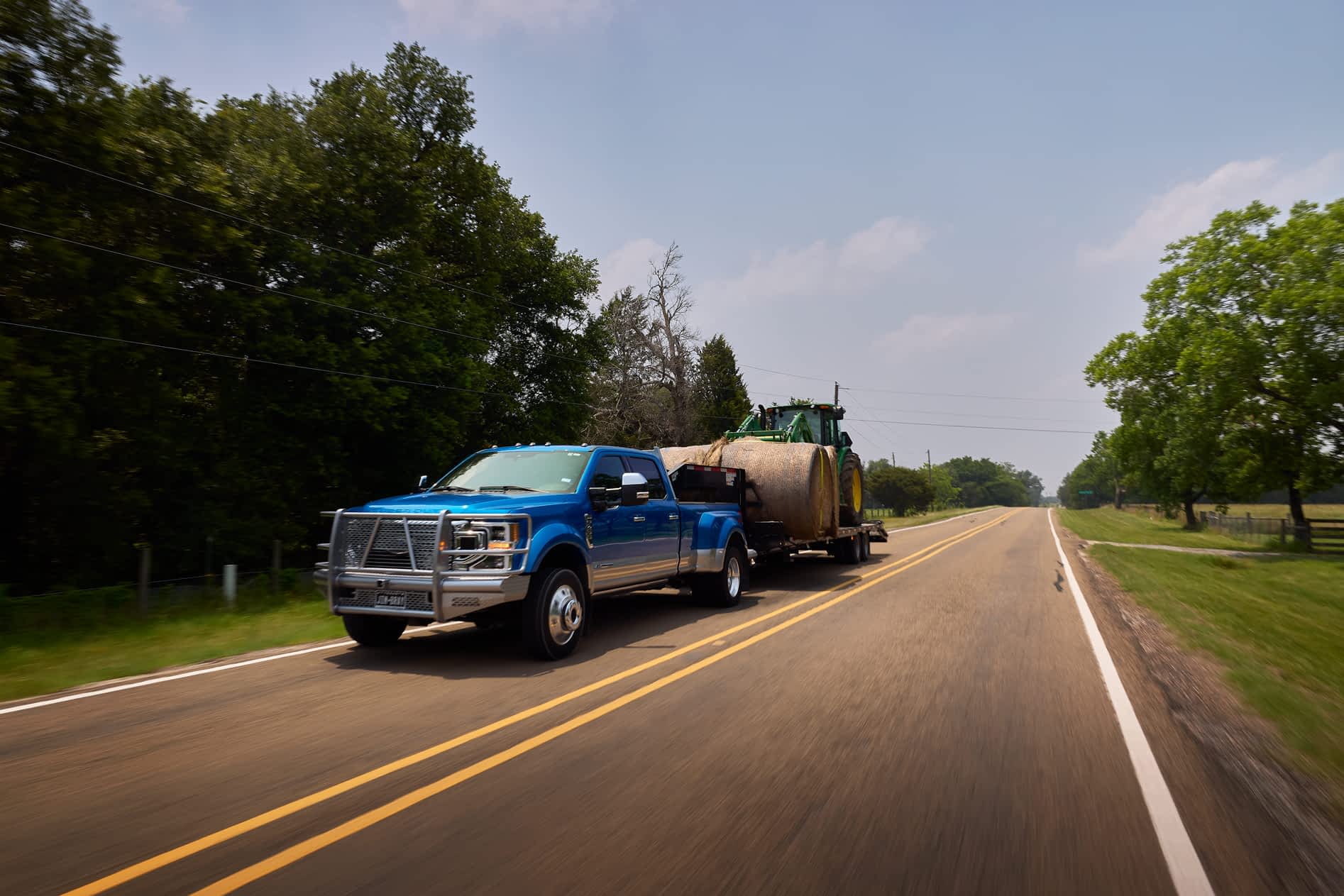 A blue dually truck pulls a Texas Pride Trailers trailer loaded with a hay bale and a green tractor on a road.