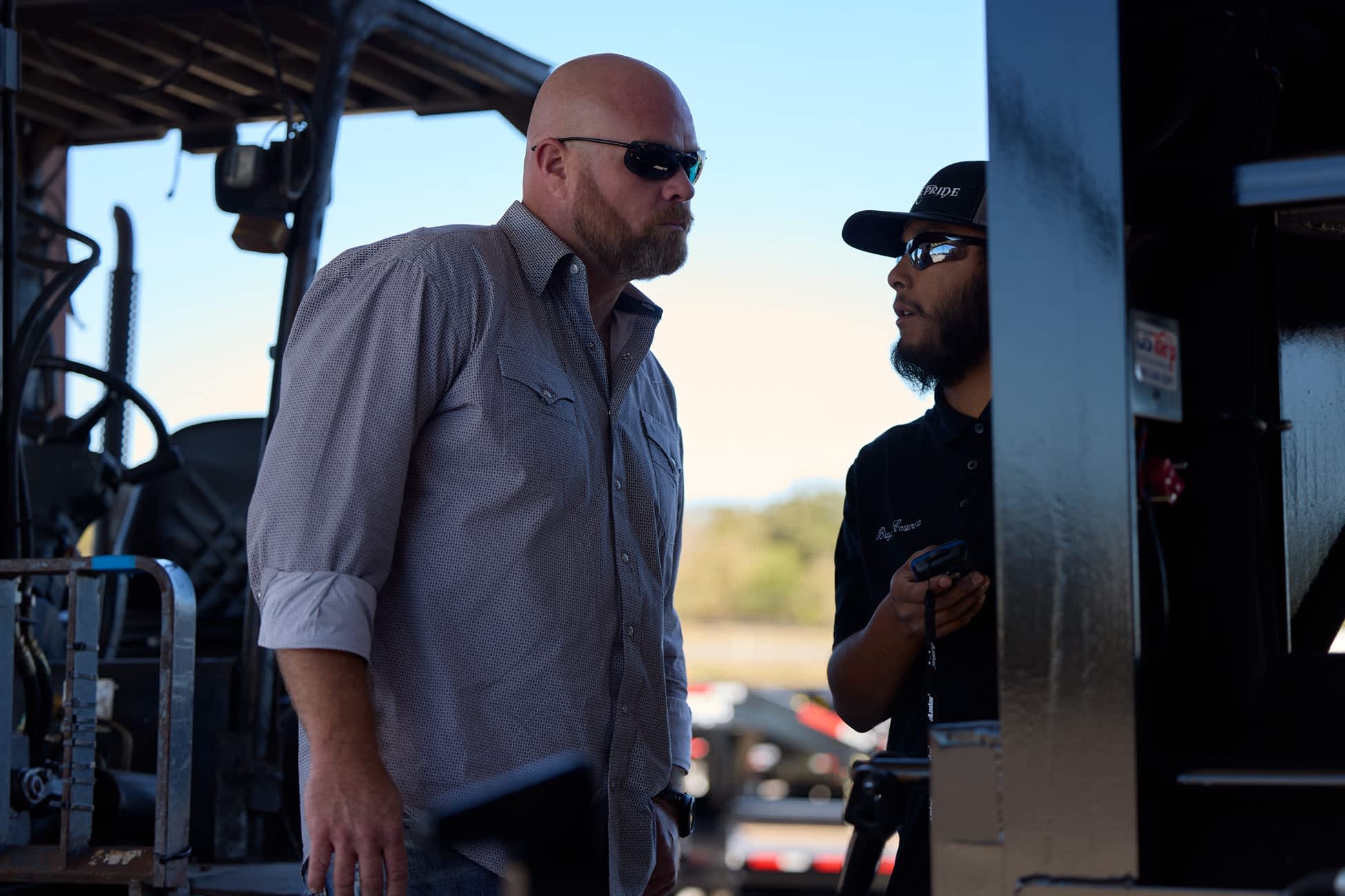 Here is the screen reader-friendly ALT text for the image: Two men talk outdoors at a location with Texas Pride Trailers in the background.