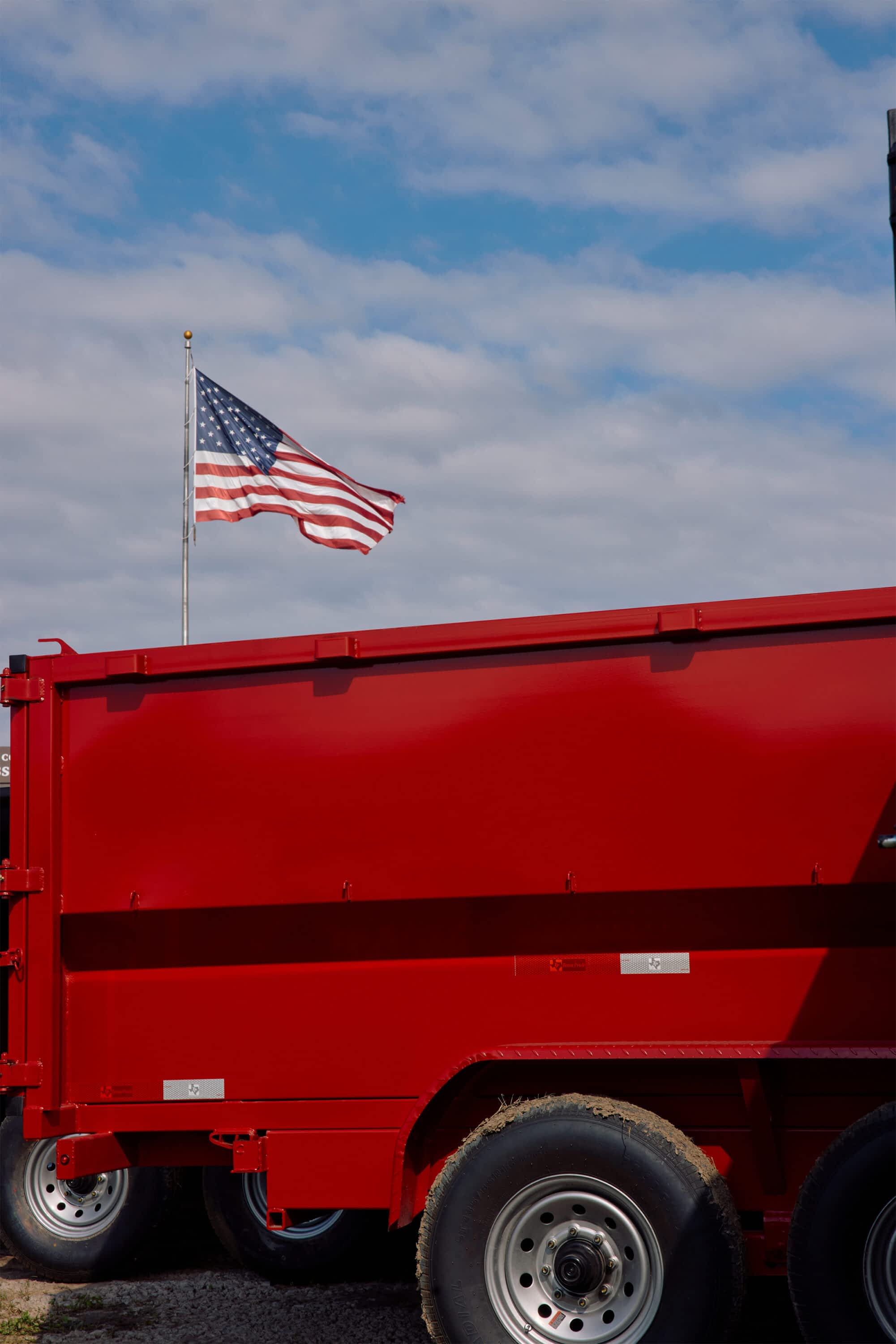 Red Texas Pride dump trailer parked outdoors with an American flag waving in the background.