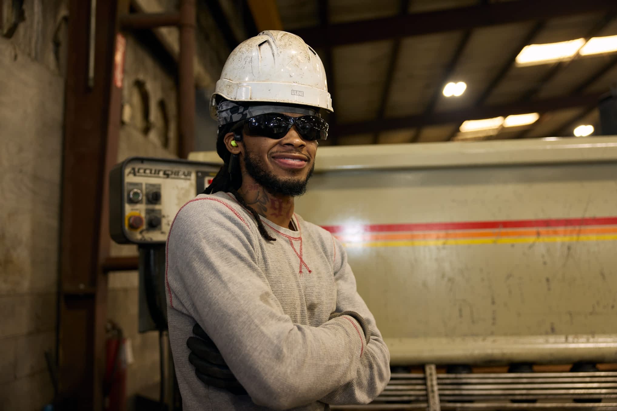 A worker in a hard hat, safety glasses, and gloves stands smiling with crossed arms in a Texas Pride Trailers manufacturing facility.
