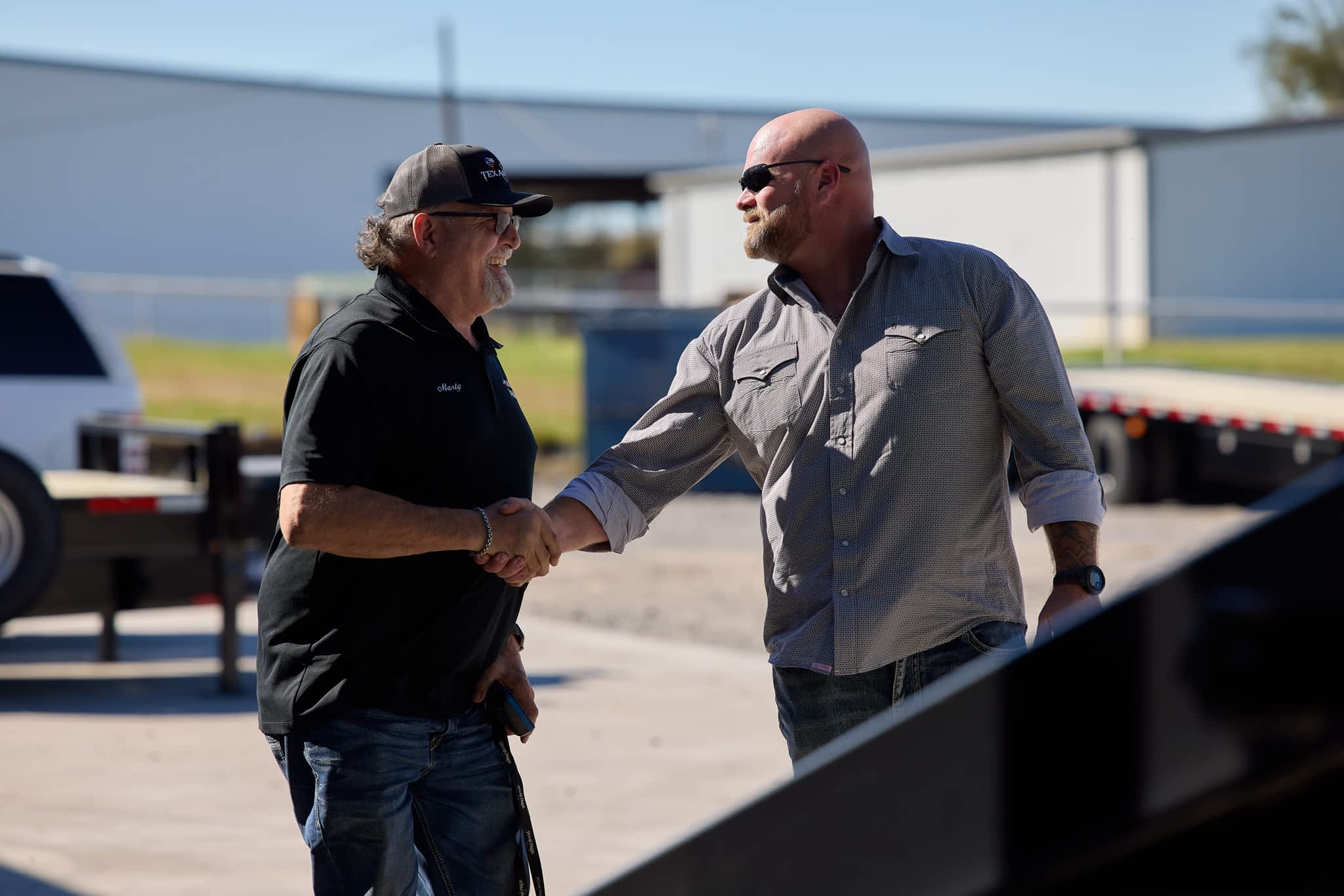 Two men shake hands outdoors near a Texas Pride Trailers trailer and buildings, suggesting a business interaction.