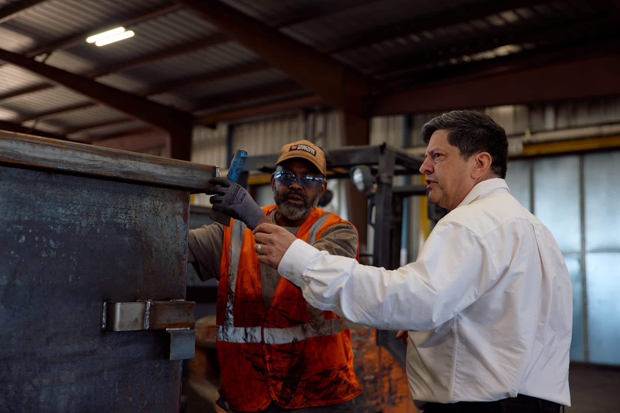 Two men inspect a metal component in a factory, likely part of a Texas Pride Trailers manufacturing process. One man in a safety vest holds a tool, while another in a shirt and tie points.