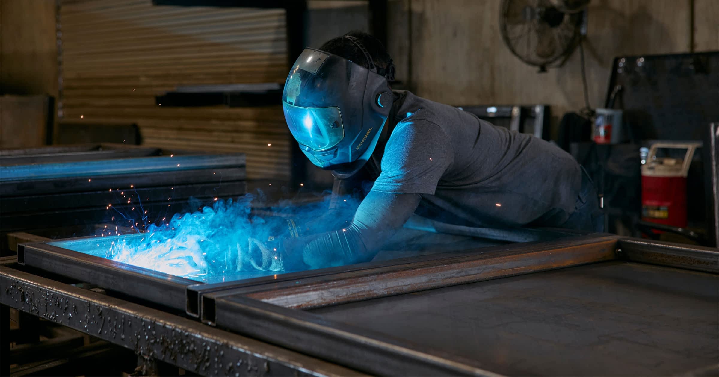 A welder works on a trailer frame with glowing blue sparks flying.