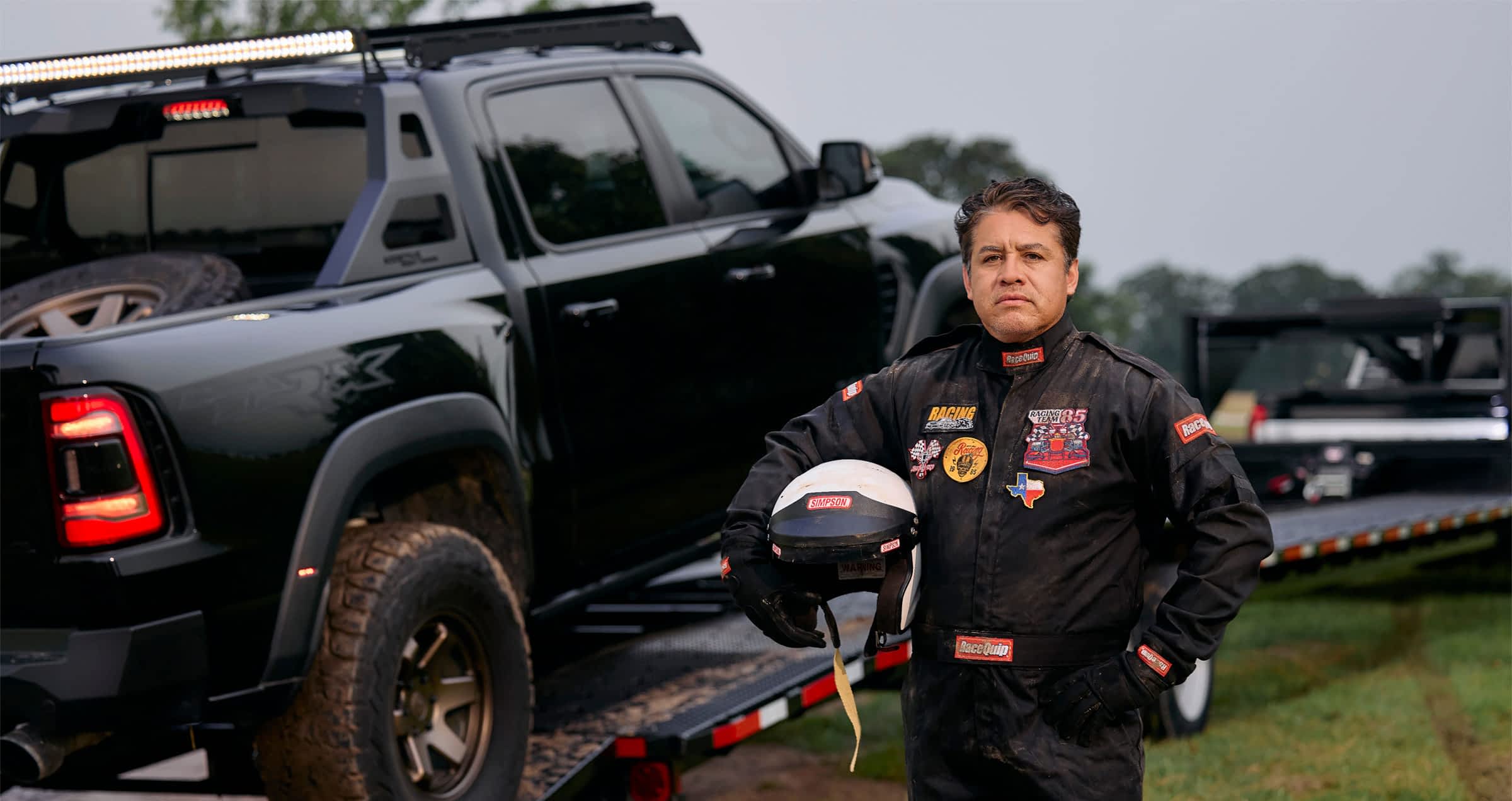 Racing driver in full gear standing beside a Texas Pride car hauler trailer loaded with an off-road vehicle.