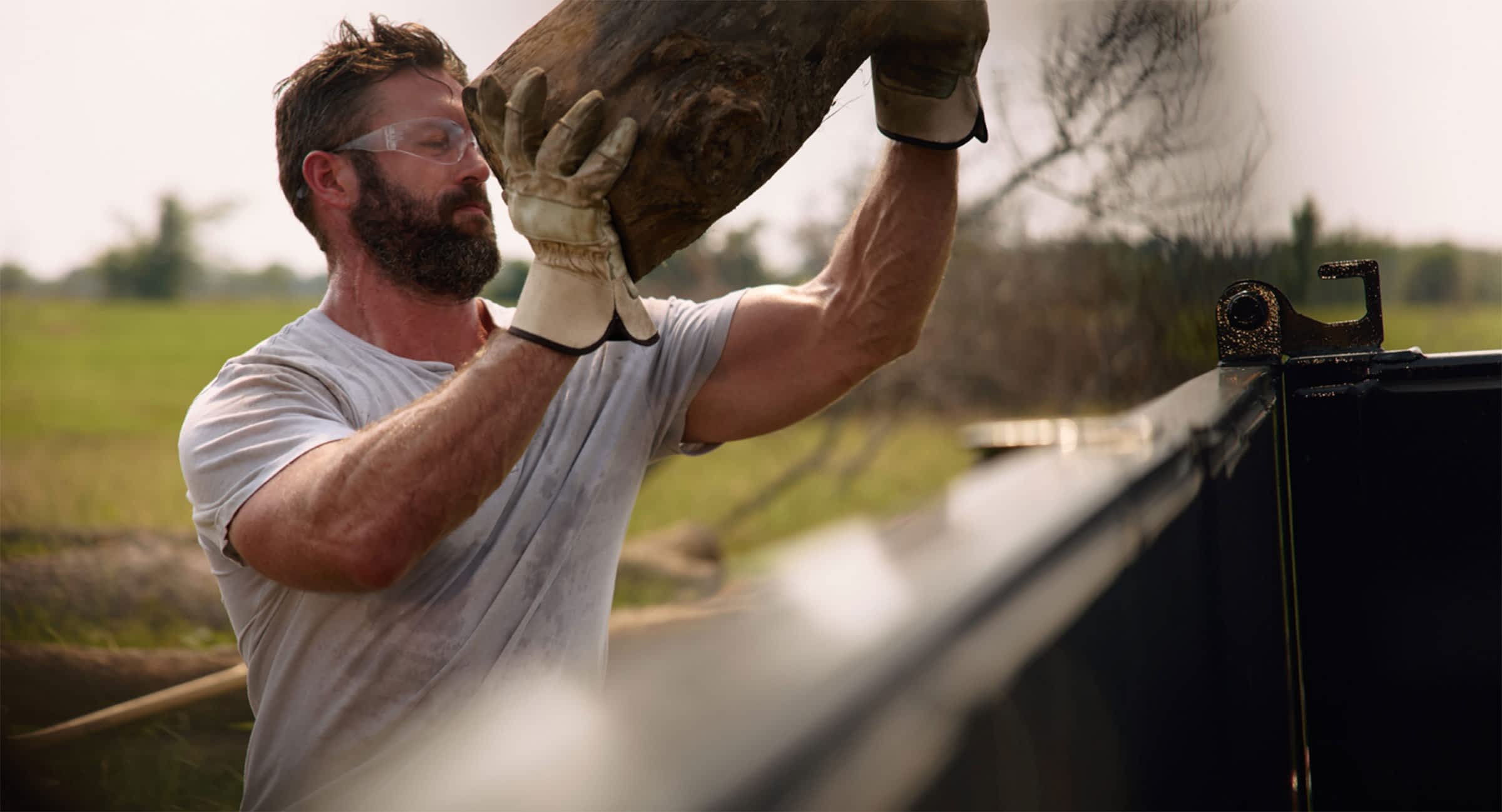 A man in safety glasses and gloves lifts a log to throw into a Texas Pride Trailers dump trailer