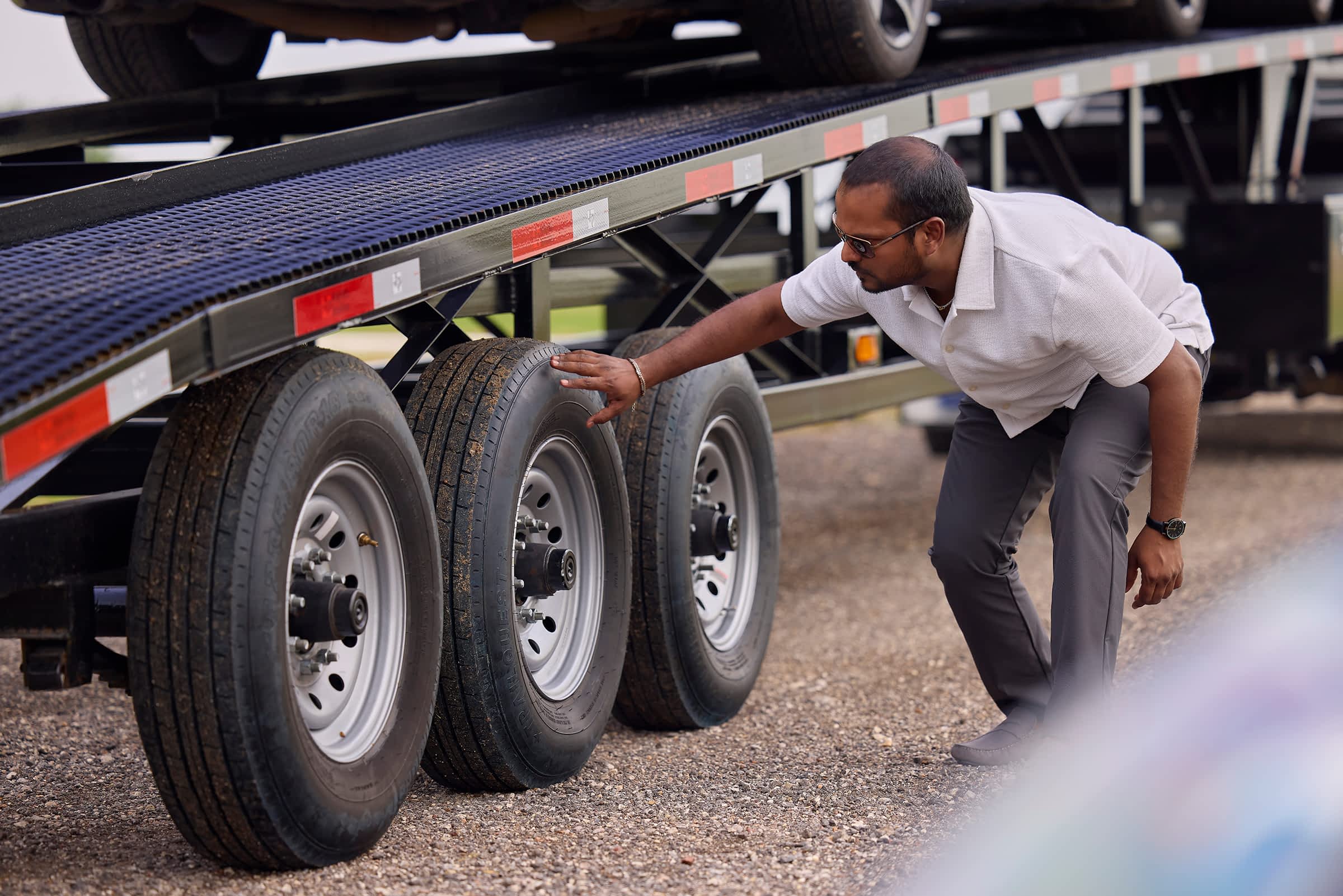 Man inspecting the triple axle wheels of a Texas Pride trailer parked on gravel.