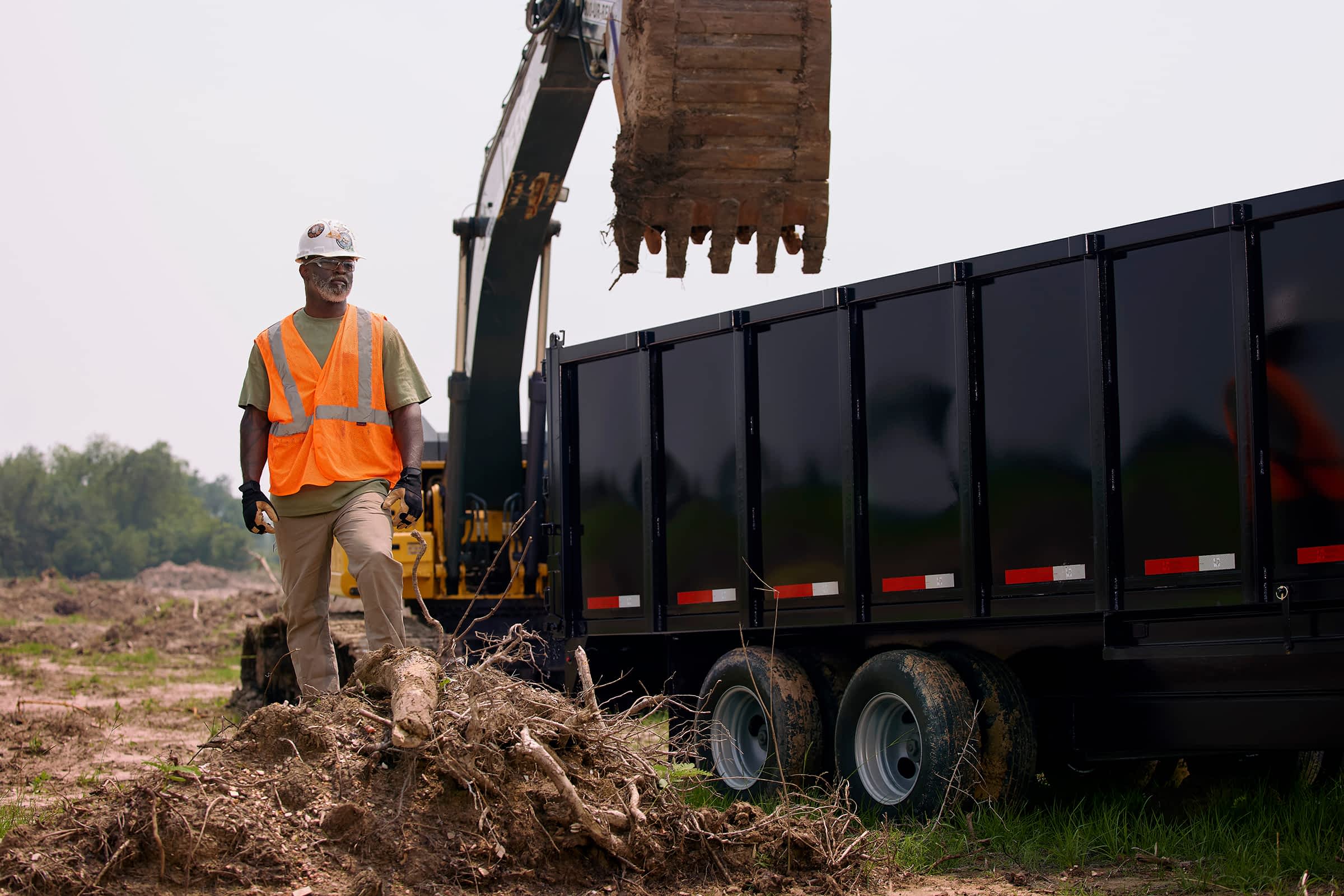 Worker supervising soil loading into a Texas Pride trailer with an excavator on a construction site.