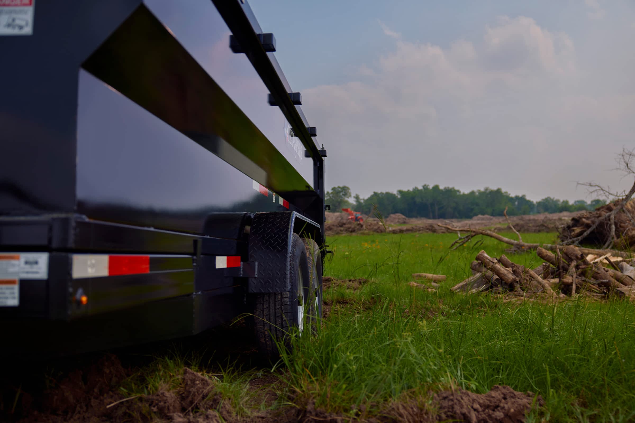 A black dump trailer sits on a muddy patch of ground with tall green grass surrounding it. In the background, a partly cloudy sky meets a line of trees.