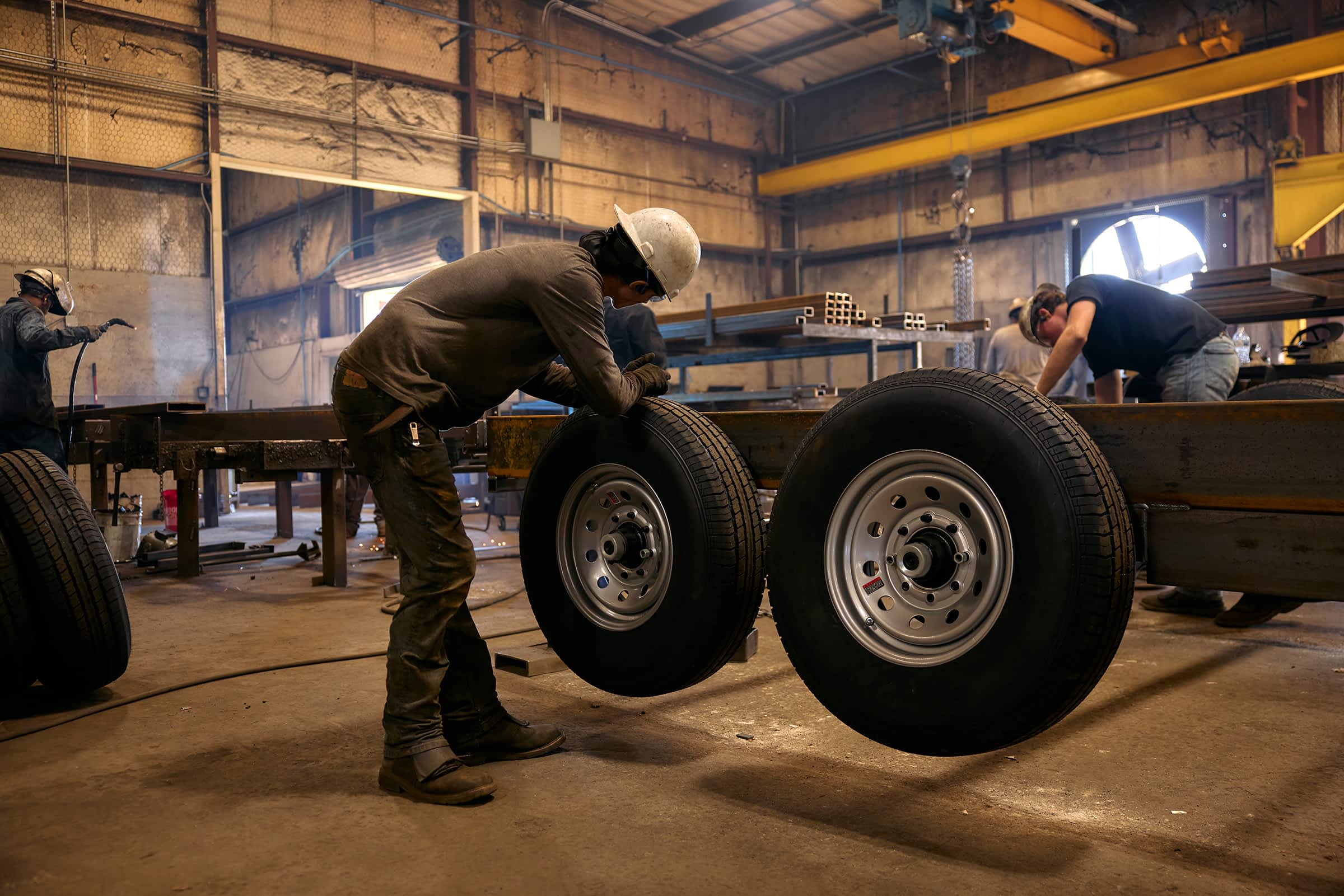 Two workers in a factory installing wheels onto a metal trailer frame, wearing safety gear and working in a large industrial workshop.