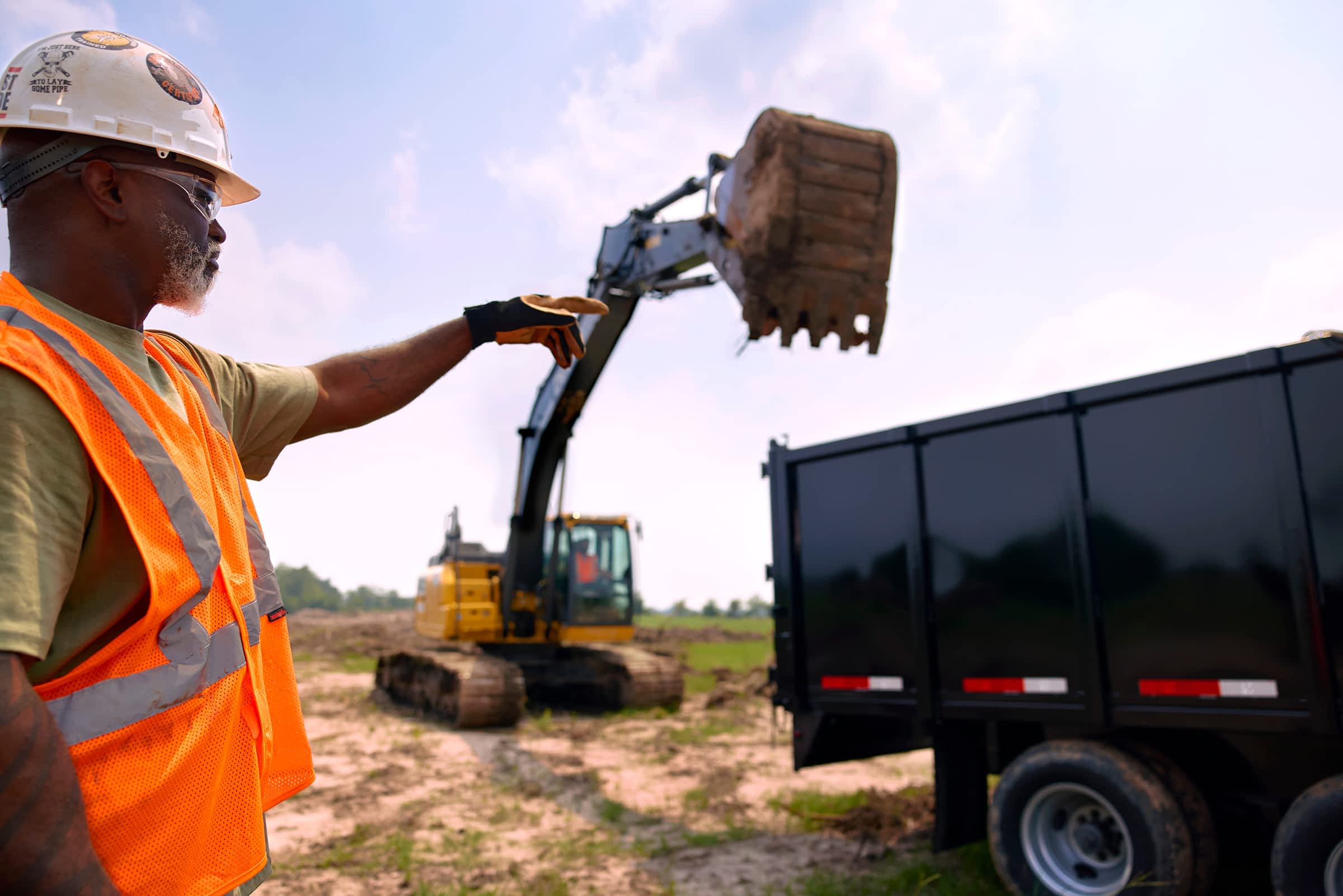Construction worker directing an excavator loading debris into a Texas Pride dump trailer on a cleared jobsite.