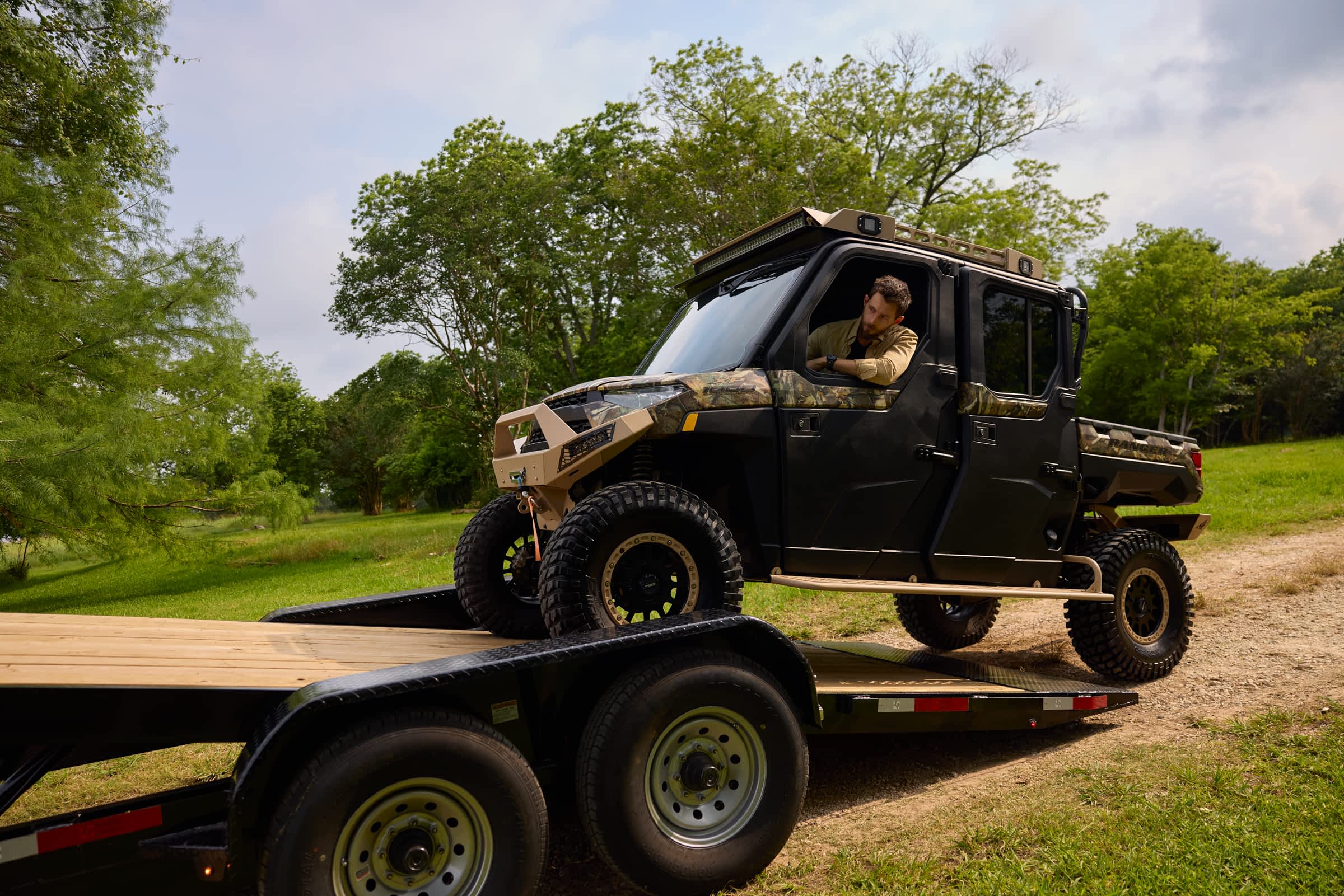 A man drives a tan-and-black off-road utility side-by-side up a Texas Pride gravity tilt trailer with a wood deck, positioned on a grassy path surrounded by trees—demonstrating easy equipment loading in a rugged outdoor setting.