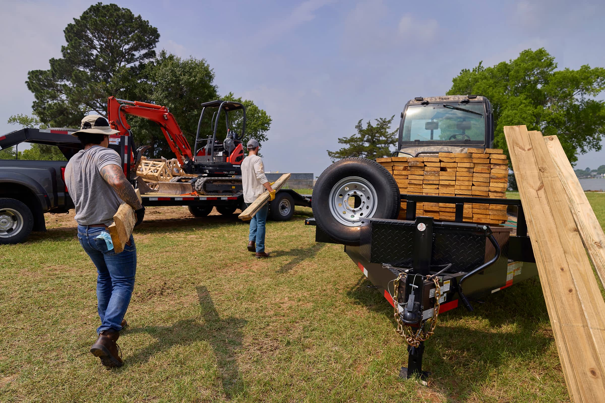 Two workers carrying lumber between Texas Pride trailers loaded with timber and construction equipment at a rural jobsite.