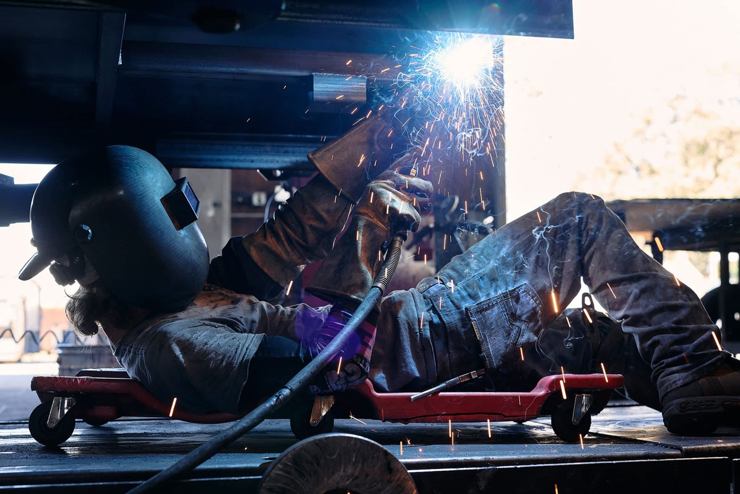 Welder lying on a creeper under a trailer frame, producing sparks while welding, wearing full protective gear including helmet and gloves.
