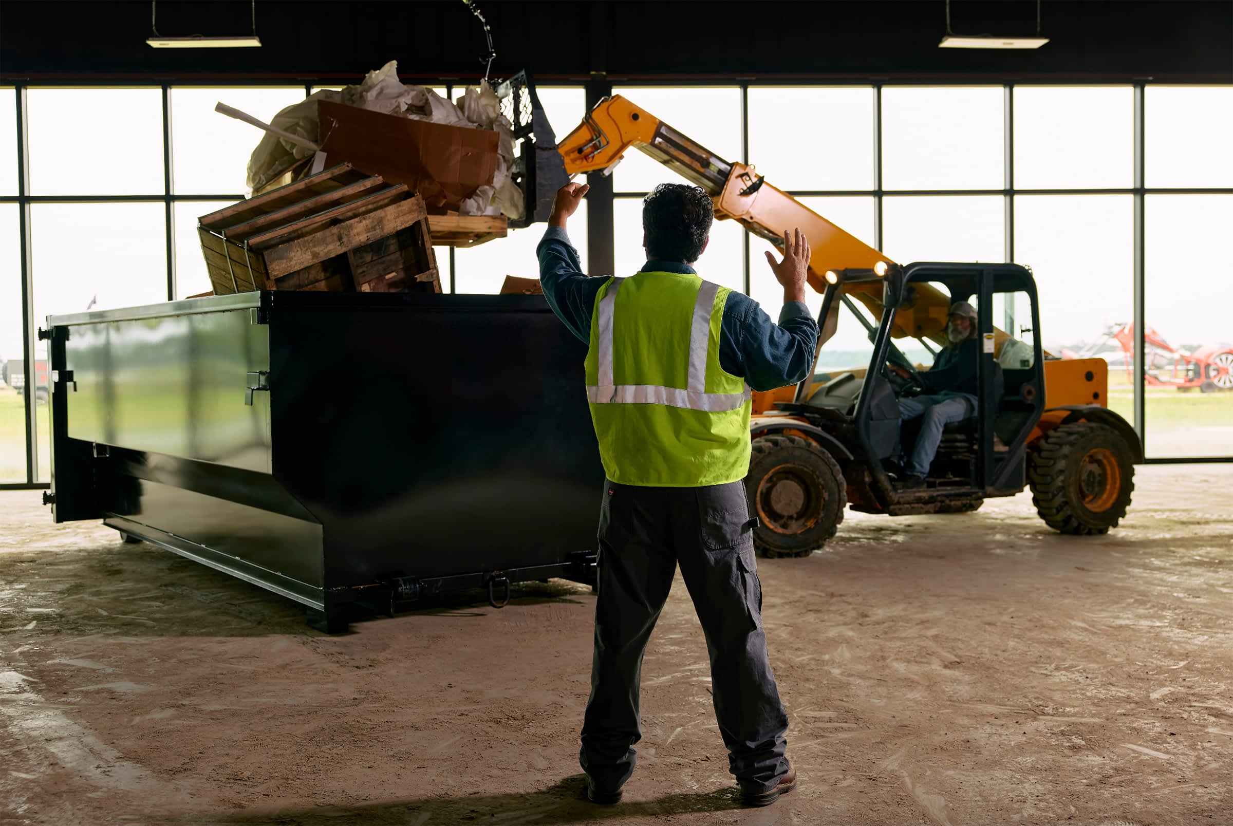 Worker guiding a telehandler as it loads debris into a Texas Pride roll-off dumpster inside a large industrial building.