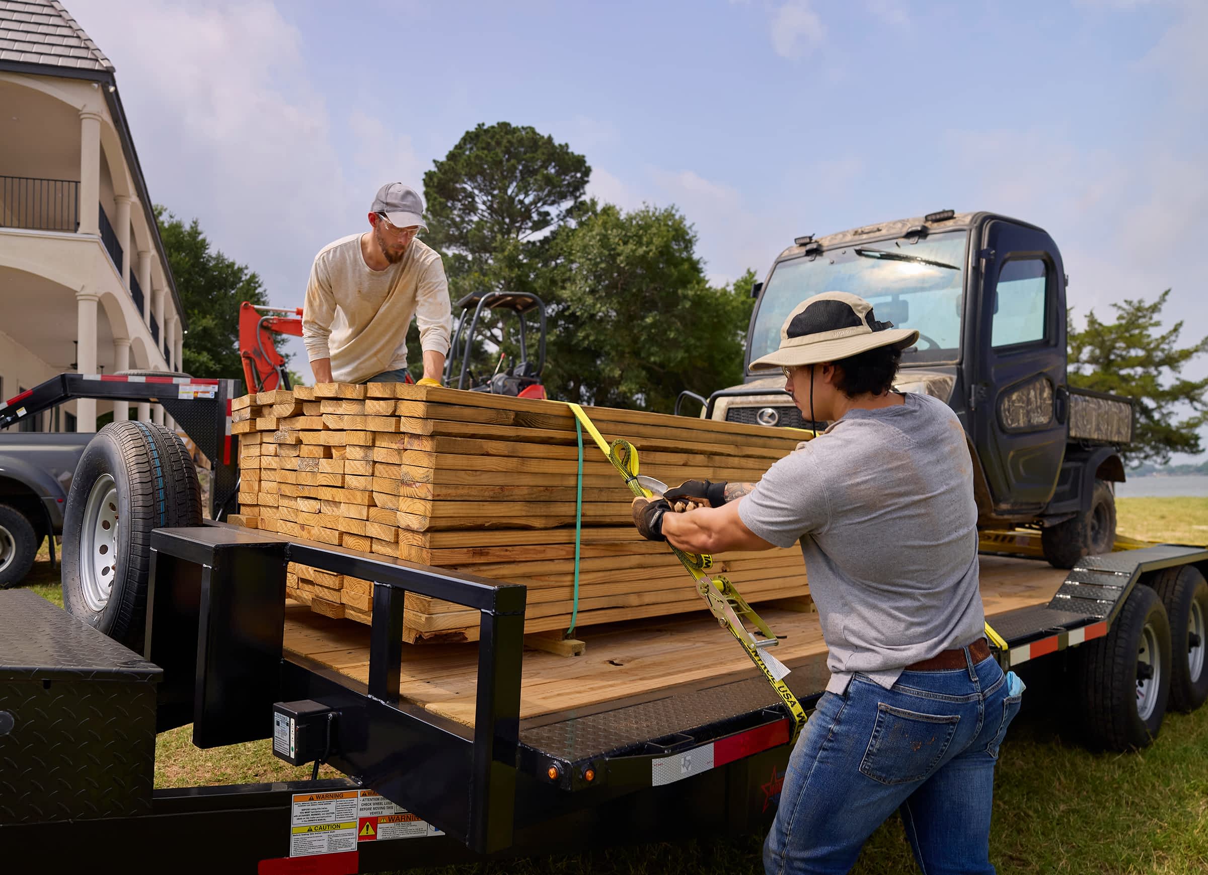 Two men securing a stack of lumber onto a Texas Pride flatbed trailer using ratchet straps.