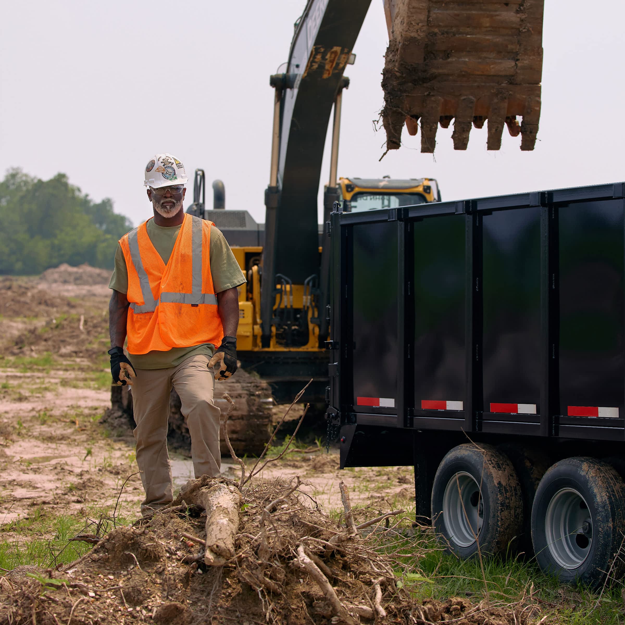 A man in an orange safety vest and white hard hat at a work site next to an excavator and dump trailer.