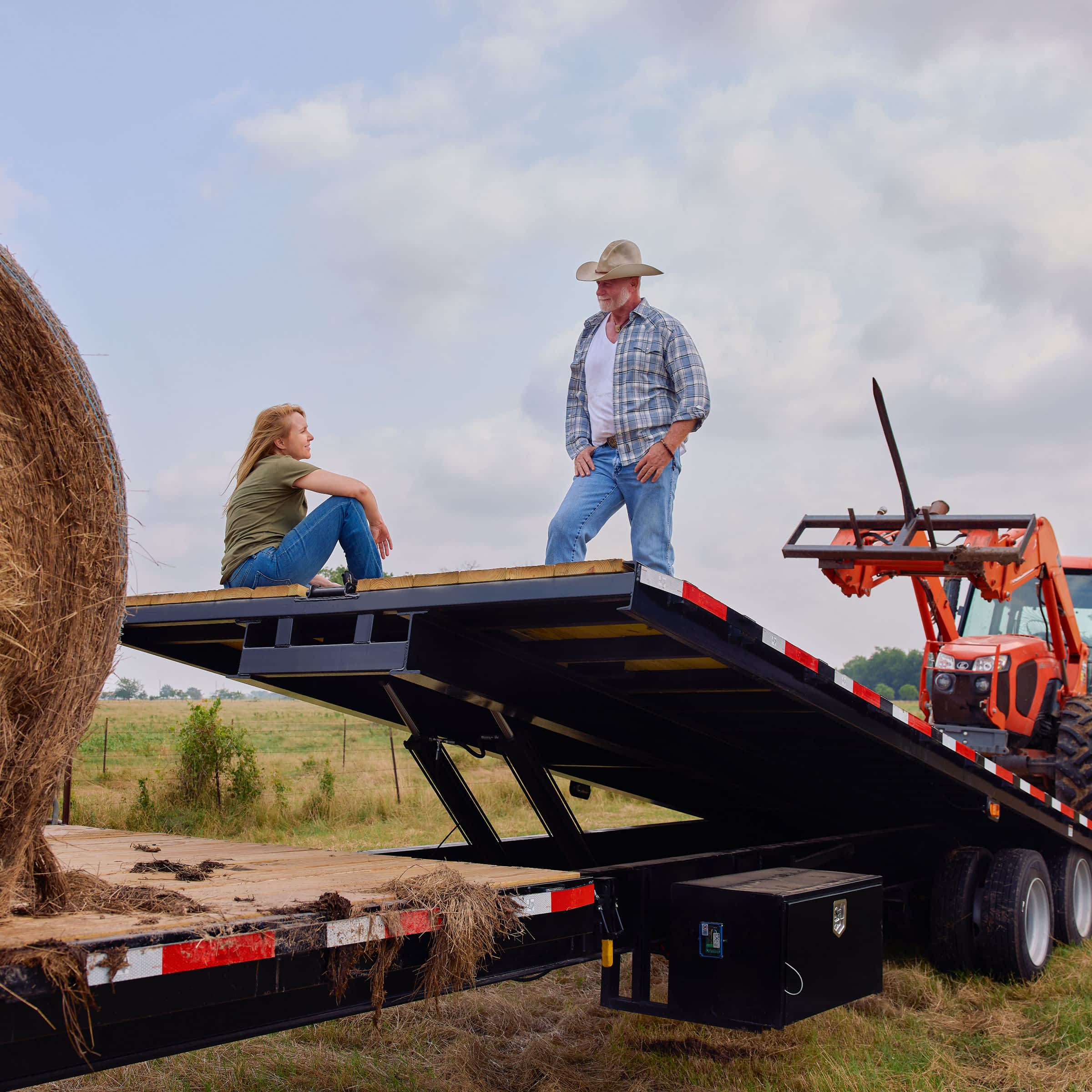 A ranch looking scene with a man and woman taking a break on a gravity tilt trailer near a tractor and bail of hay.